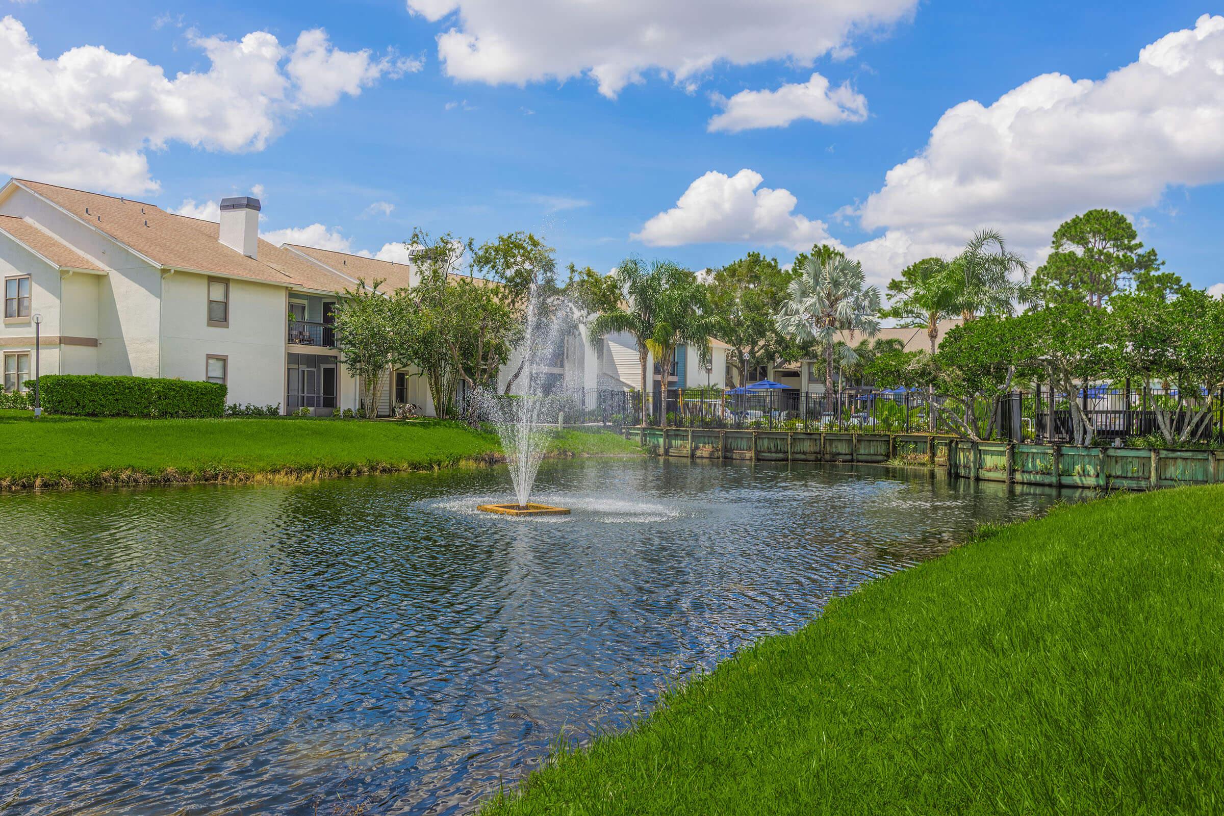 A serene lake scene featuring a decorative fountain in the center, surrounded by lush green grass and trees. In the background, there are residential buildings with a clear blue sky and fluffy white clouds above. The water reflects the vibrant colors of the landscape, creating a peaceful atmosphere.