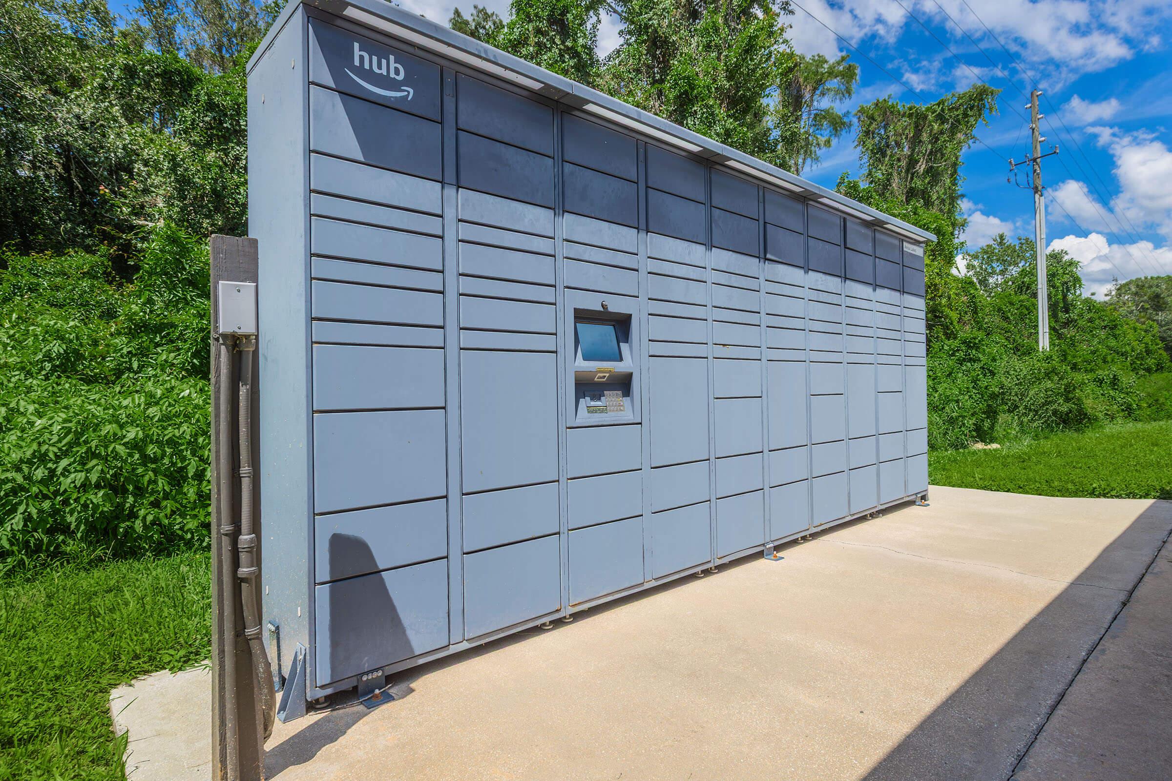 A gray Amazon delivery locker unit situated on a concrete surface, surrounded by greenery and trees under a clear blue sky. The locker features several compartments for packages, with a retrieval slot visible at the front.