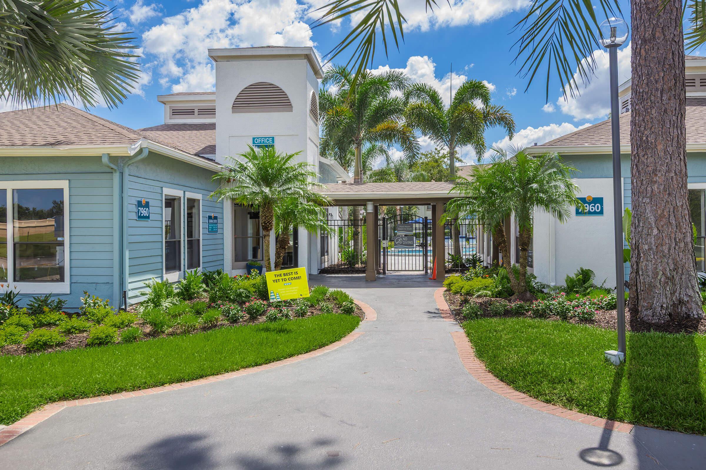 A welcoming entrance to a light blue office building surrounded by lush greenery and palm trees. The pathway leads to a gated entrance, with bright yellow signage nearby. The sky is clear with scattered clouds, adding to the inviting atmosphere of the location.