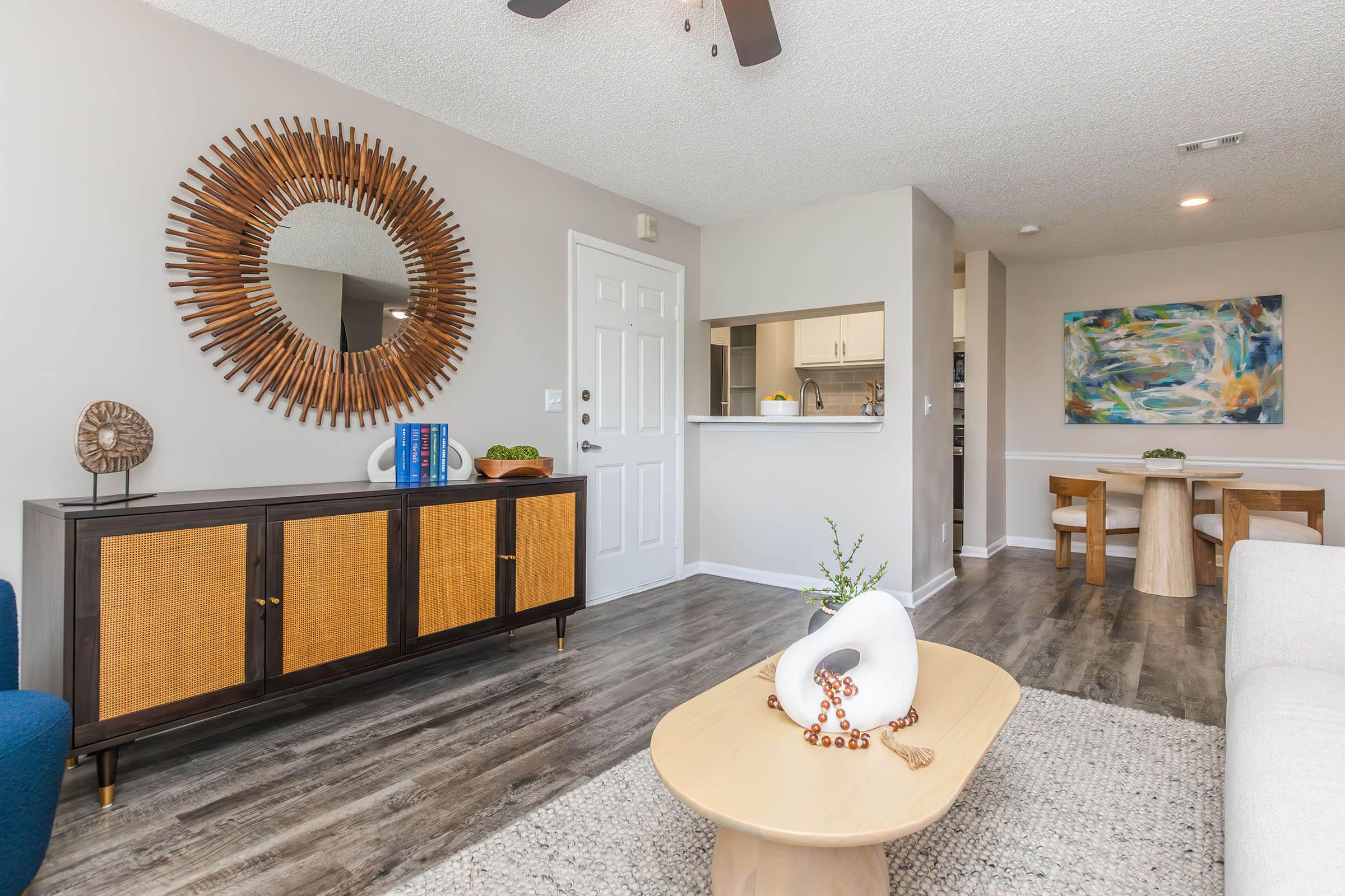 A modern living room featuring a stylish round mirror on the wall, a wooden sideboard with woven accents, a small coffee table with decorative items, and light-colored furniture. The space has a neutral color palette, wooden floors, and a dining area in the background with a colorful piece of artwork.