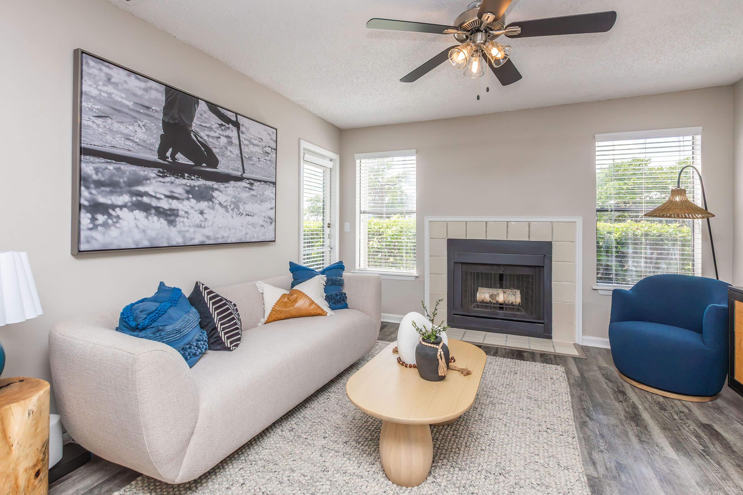 A modern living room featuring a light-colored sofa with decorative pillows, a round wooden coffee table, and a stylish blue armchair. There's a fireplace with a minimalist design, large windows allowing natural light, and a framed black-and-white photo of a person surfboarding on the wall.