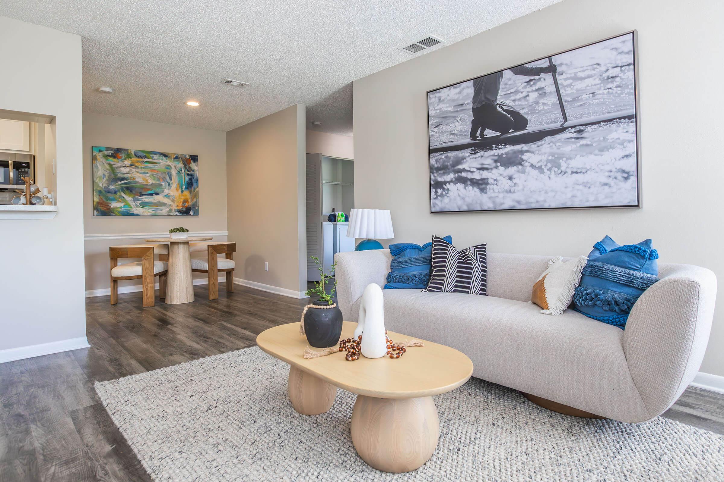 A modern living room featuring a light-colored sofa with decorative pillows, a round wooden coffee table, and a large wall art piece depicting a person paddling. The room has light-colored walls, a textured rug, and a dining area in the background with a table and chairs, creating a cozy and inviting atmosphere.