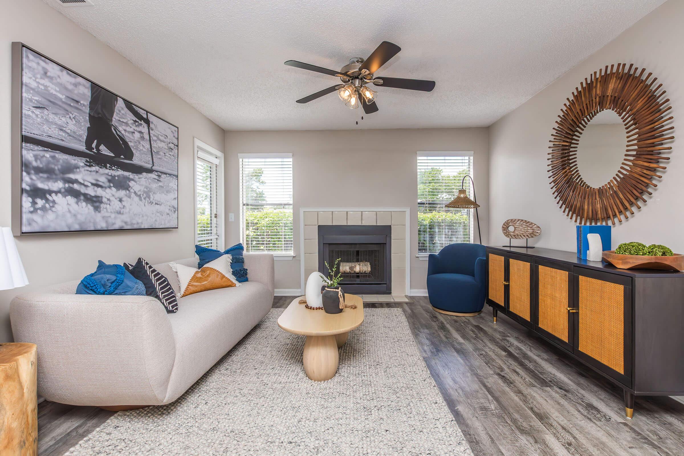 A cozy living room featuring a light gray sofa with blue and orange pillows, a wooden coffee table, and a stylish sideboard. The space has large windows with natural light, a fireplace, and modern decor including a round wall mirror and a large black-and-white photograph. Hardwood flooring adds warmth to the ambience.