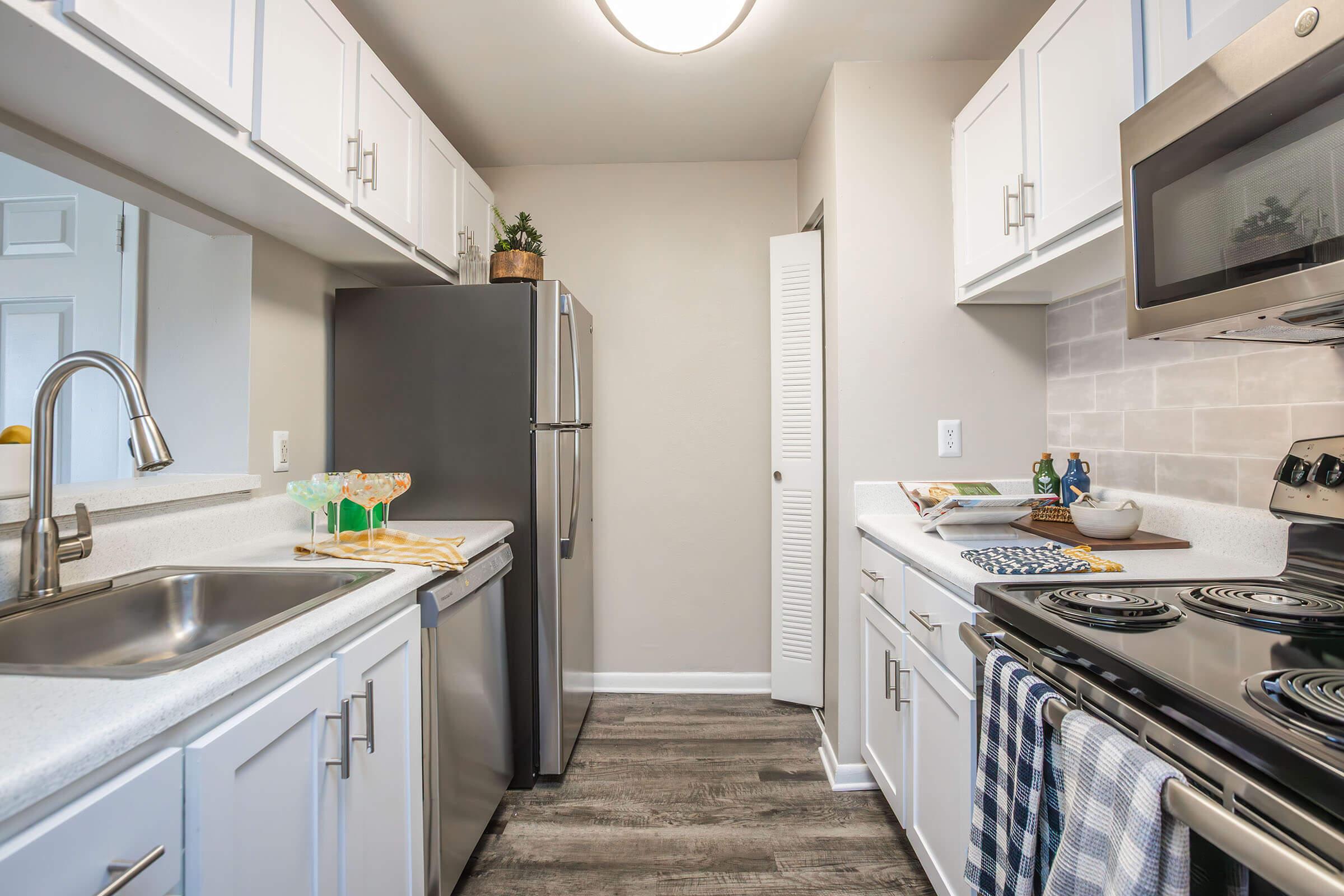 A modern kitchen with white cabinetry, stainless steel appliances, and a gray stone backsplash. The layout includes a double sink, oven, stove, and a refrigerator. Natural light illuminates the space, highlighting decorative elements like a plant and kitchen accessories on the countertop and table.