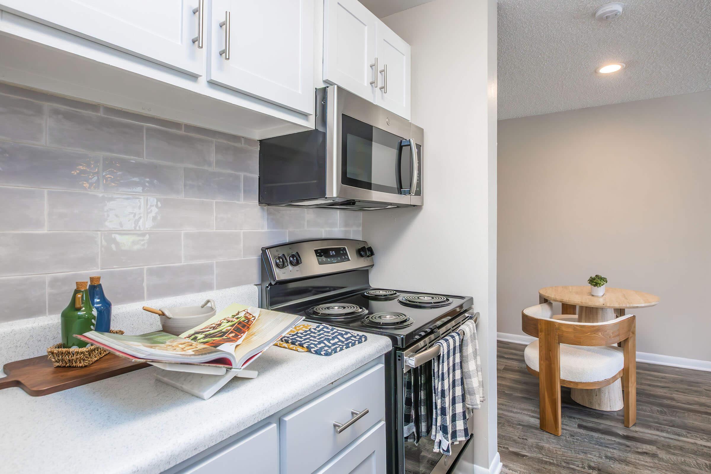 Modern kitchen featuring stainless steel appliances, including a microwave and a stovetop. A cookbook lies open on the countertop beside a wooden serving tray with bottles. The decor includes white cabinetry and light gray walls, with a small dining table visible in the background.