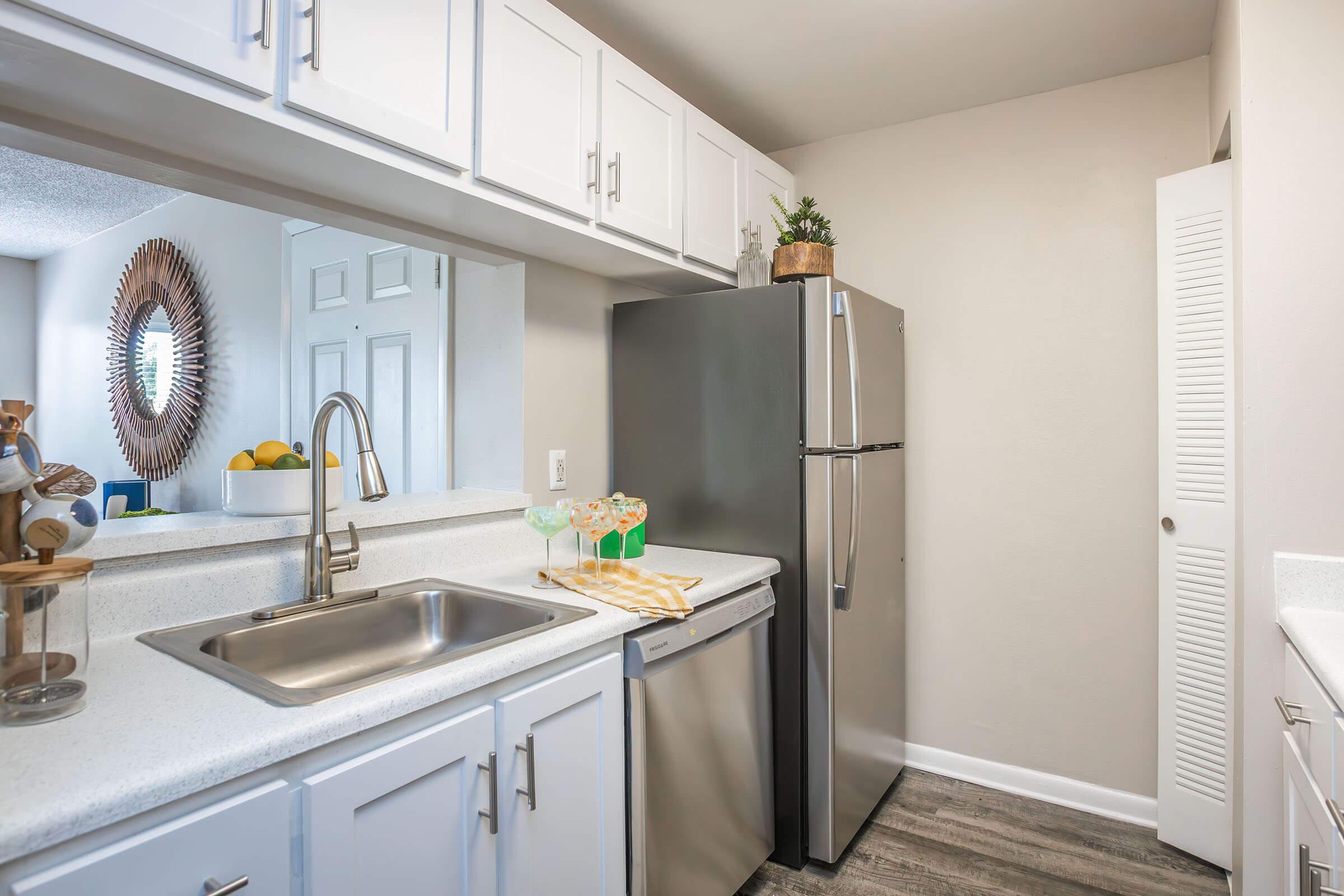 A modern kitchen featuring white cabinets, a stainless steel refrigerator, and a sink. There’s a dishwasher beside the sink, and a decorative mirror on the wall. A counter holds a tray with drinks and a small plant, while various fruits are displayed in a bowl. The flooring has a wood-like appearance.