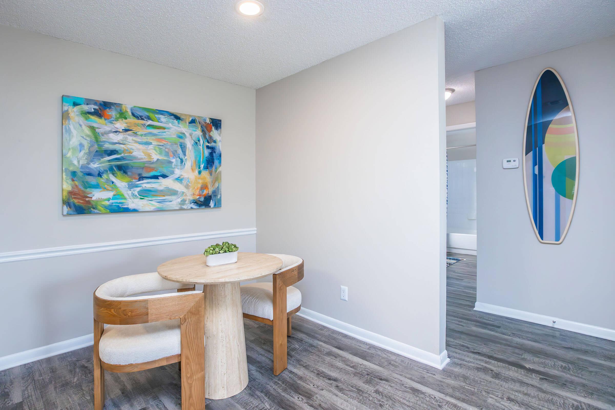A modern interior featuring a small round wooden dining table with two cushioned chairs. The wall is adorned with a colorful abstract painting, and a narrow oval mirror hangs nearby. The flooring is a light wood finish, and the walls are painted in soft neutral tones, contributing to a bright and inviting atmosphere.