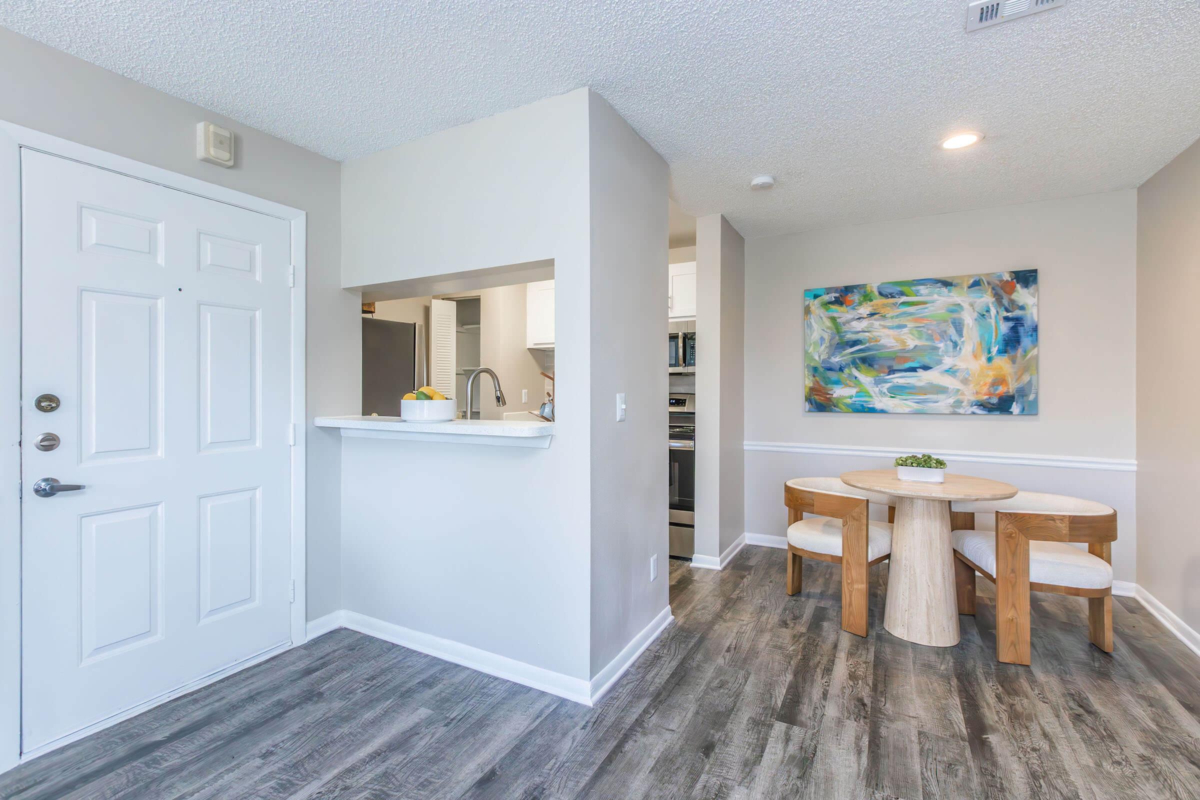 A bright and modern apartment interior featuring a small dining area with a round wooden table and two white chairs. The space includes a white kitchen with a pass-through counter and a colorful abstract painting on the wall. Light-colored walls and laminate flooring create a welcoming atmosphere.