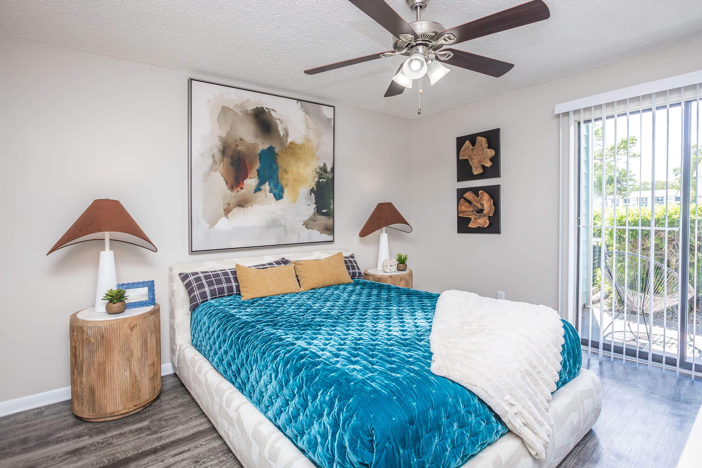 A modern bedroom featuring a large bed with a vibrant teal comforter, yellow accent pillows, and a white throw blanket. Two lamps with brown shades are on wooden side tables. A decorative wall art piece is above the bed, and abstract wooden sculptures are displayed on the wall. A ceiling fan and sliding glass door lead to an outdoor area.