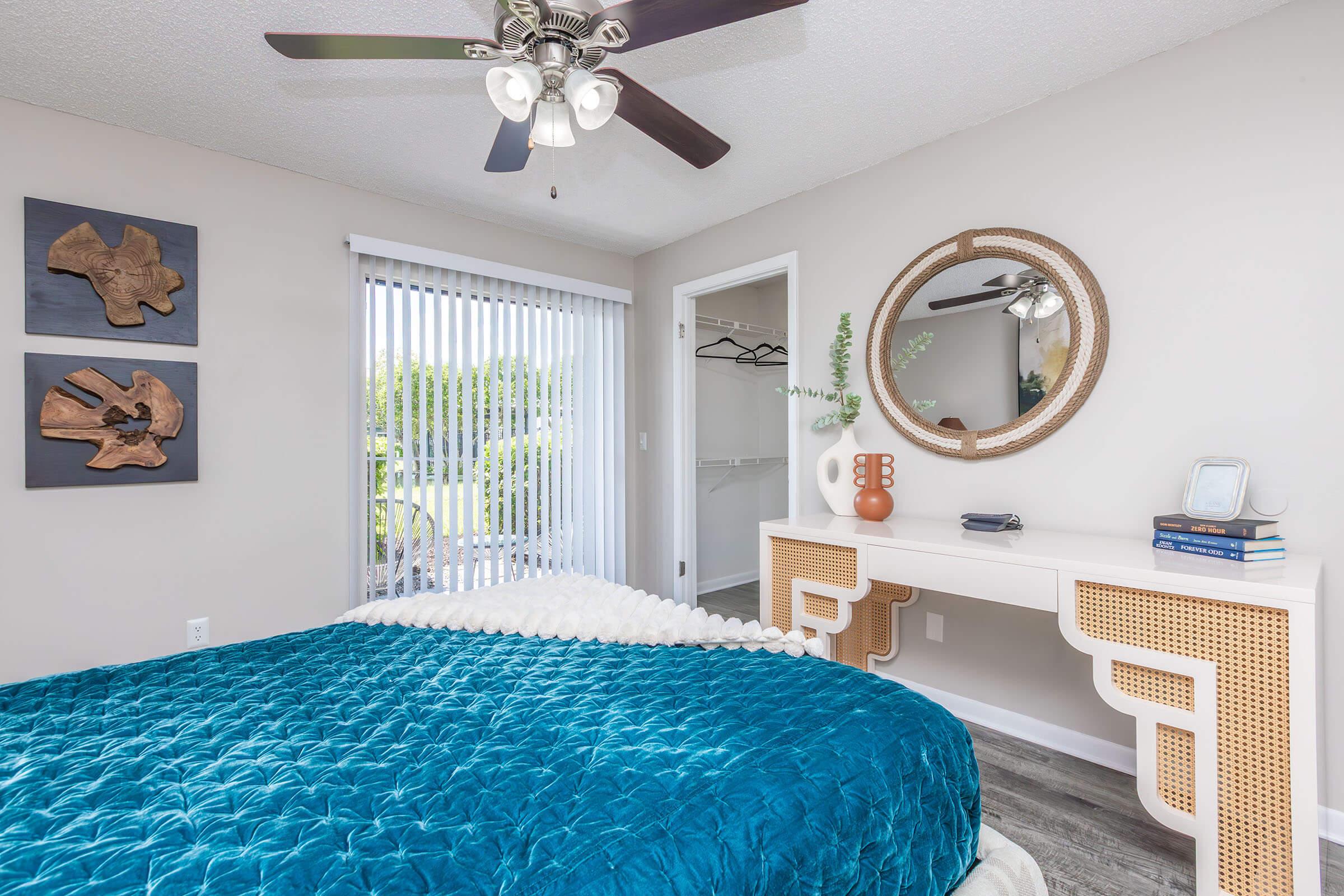 A cozy bedroom featuring a teal quilted bedspread, a stylish white dresser with circular mirror, and decorative items. The room has a ceiling fan, wooden wall art, a sliding glass door with vertical blinds, and a closet space visible in the background. Natural light brightens the inviting atmosphere.