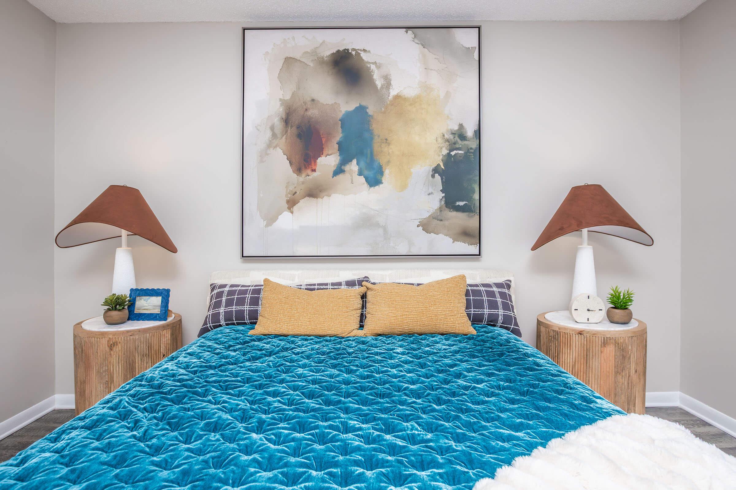 A modern bedroom featuring a large abstract painting above a stylish bed with a bright blue quilt. The bed has two yellow pillows and is flanked by wooden side tables with brown lamps and small plants. The walls are light-colored, and the room has a contemporary aesthetic.