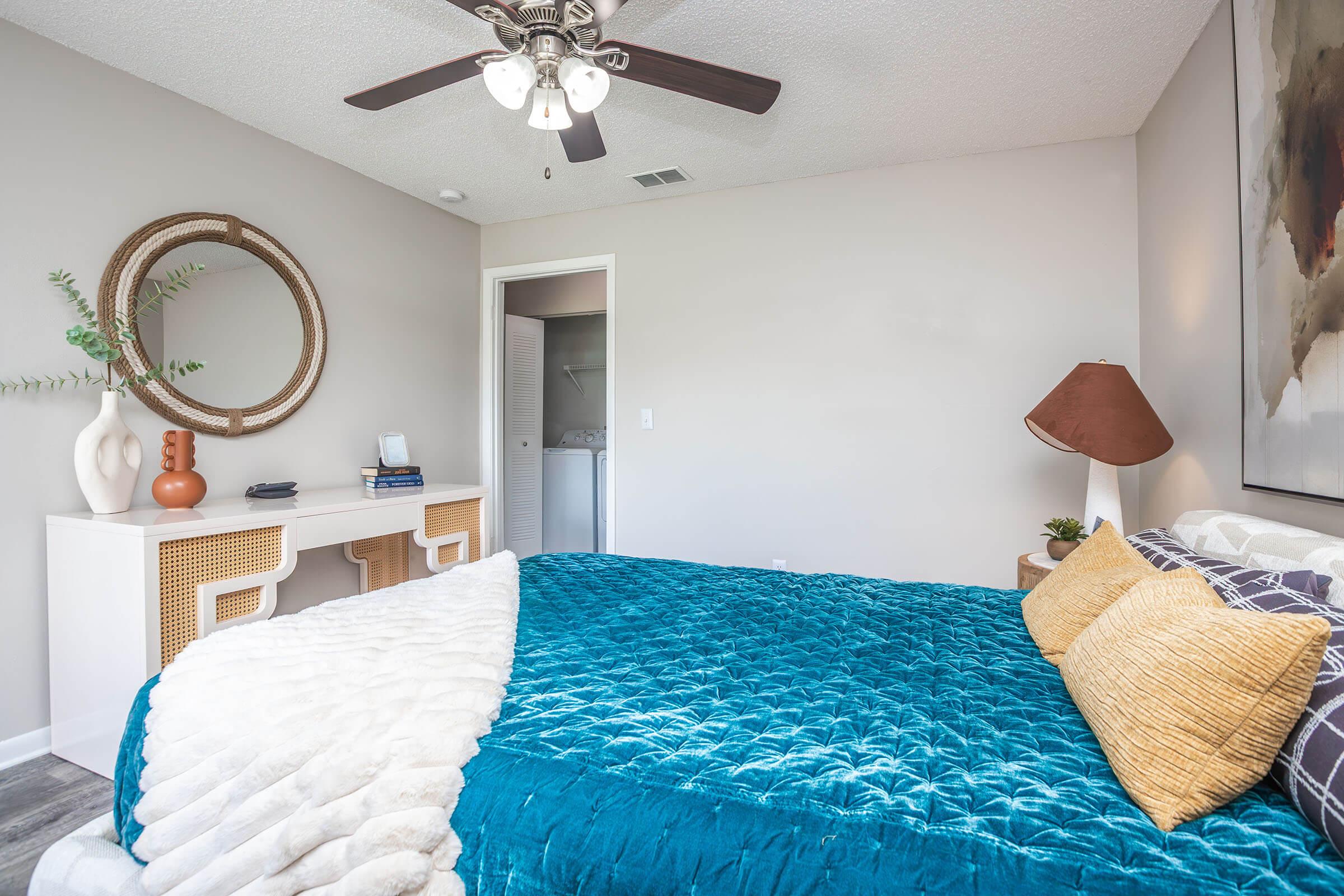A cozy bedroom featuring a queen-sized bed with a teal quilt and decorative pillows. The room includes a modern bedside lamp, a large circular mirror above a white dresser, and a small desk area. Natural light comes through an open door leading to a bathroom, enhancing the inviting atmosphere.