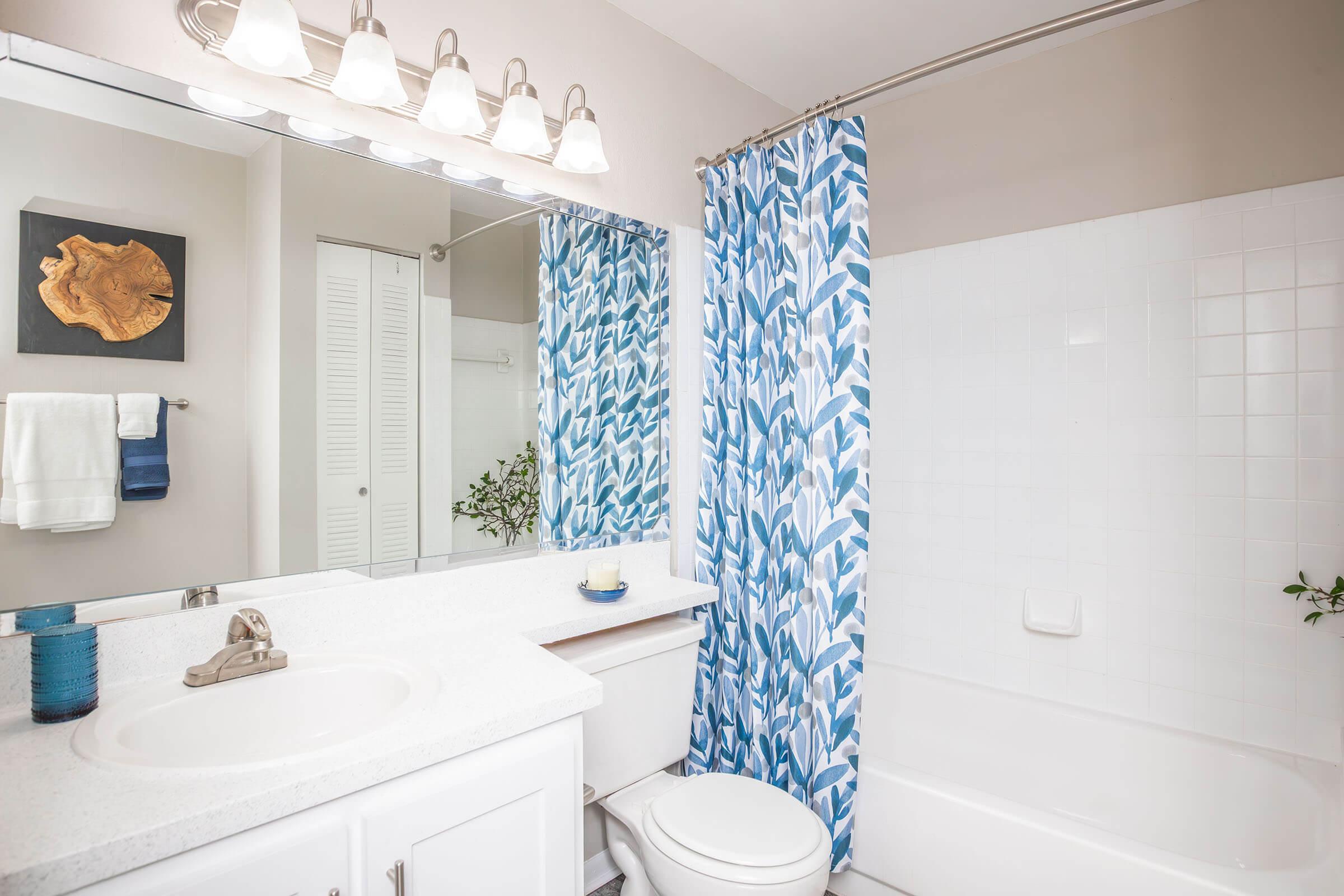 A modern bathroom featuring a white countertop sink, a large mirror, and a bathtub with a white tiled wall. The space includes a blue and white patterned shower curtain, a decorative wooden wall art piece, and light fixtures above the mirror, creating a bright and clean ambiance.