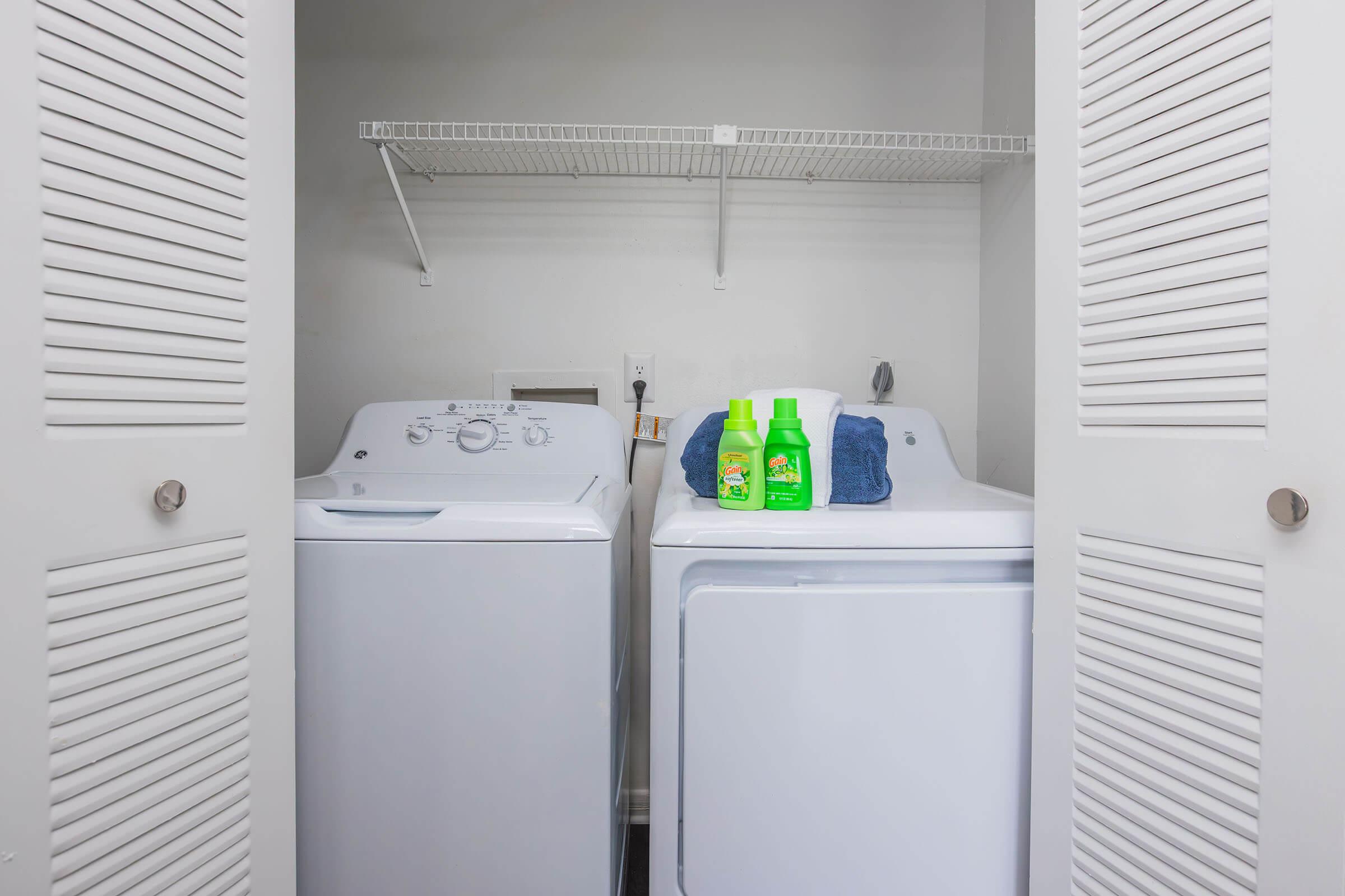 A laundry room with a white washing machine and a white dryer side by side. Above them is a shelf holding two bottles of laundry detergent and a folded blue towel. The room is enclosed by two louvered doors, creating a neat and organized appearance.