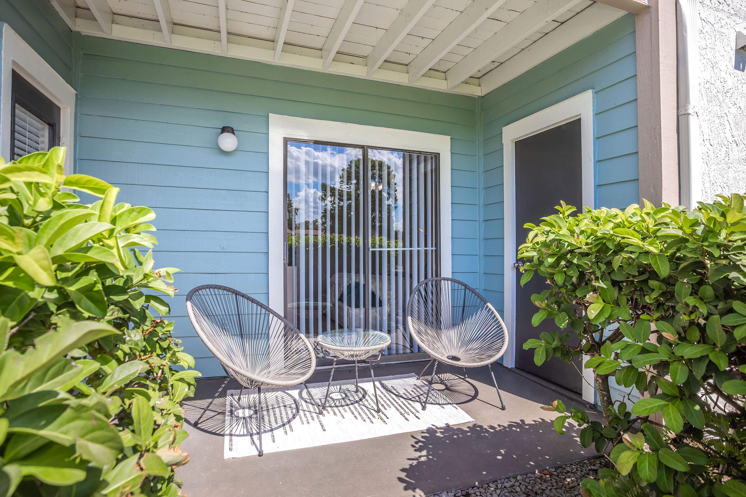 A cozy outdoor patio area with two modern chairs and a small table, framed by green shrubs. The walls are painted in a soothing light blue color, and there is a sliding glass door with vertical blinds. Daylight illuminates the space, enhancing the inviting atmosphere.