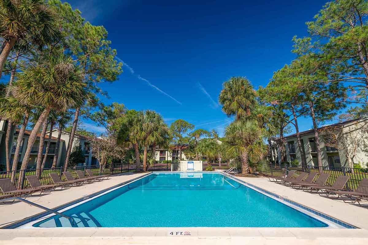 A clear blue swimming pool surrounded by palm trees and lounge chairs. The scene features bright blue skies with a few clouds, creating a relaxing atmosphere. There are several buildings in the background, adding to the resort-like setting.