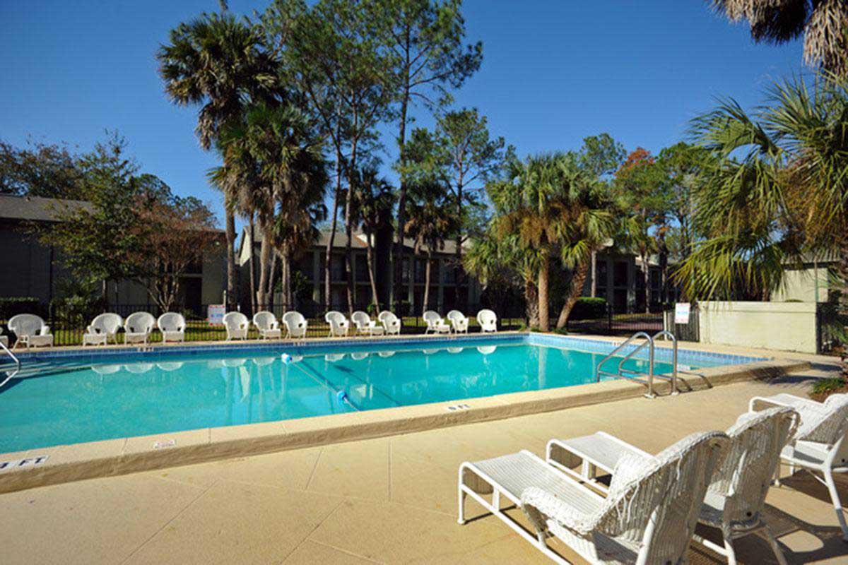 A clear blue swimming pool surrounded by palm trees and lounge chairs. The sun is shining, and the sky is bright blue. A fence lines the pool area, with a few trees in the background. The setting appears clean and inviting, perfect for relaxation and leisure.