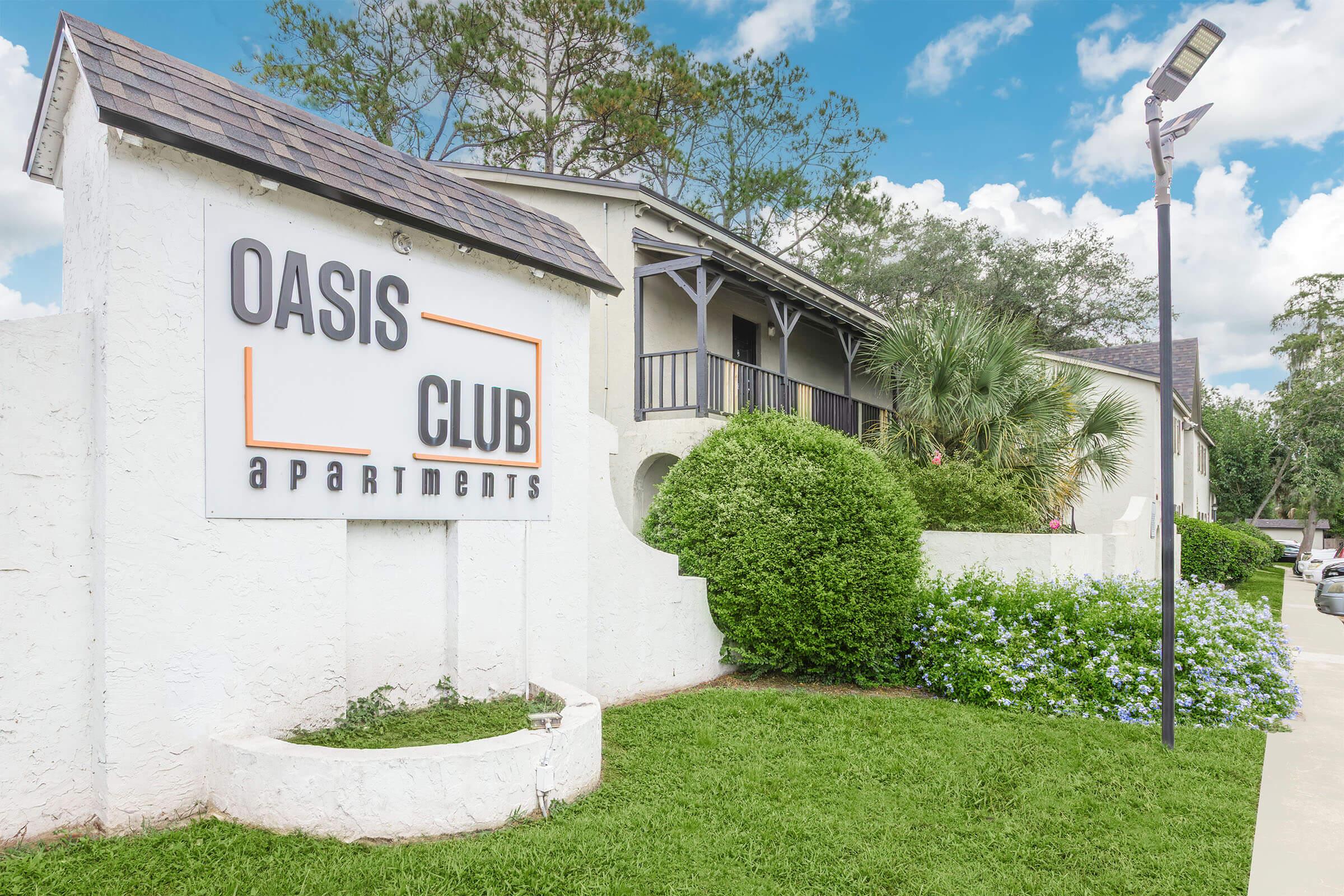 Sign for Oasis Club Apartments with a white exterior wall and lush green landscaping. The building features balconies and is surrounded by trees and flowering shrubs under a bright blue sky with scattered clouds. A streetlight stands nearby.