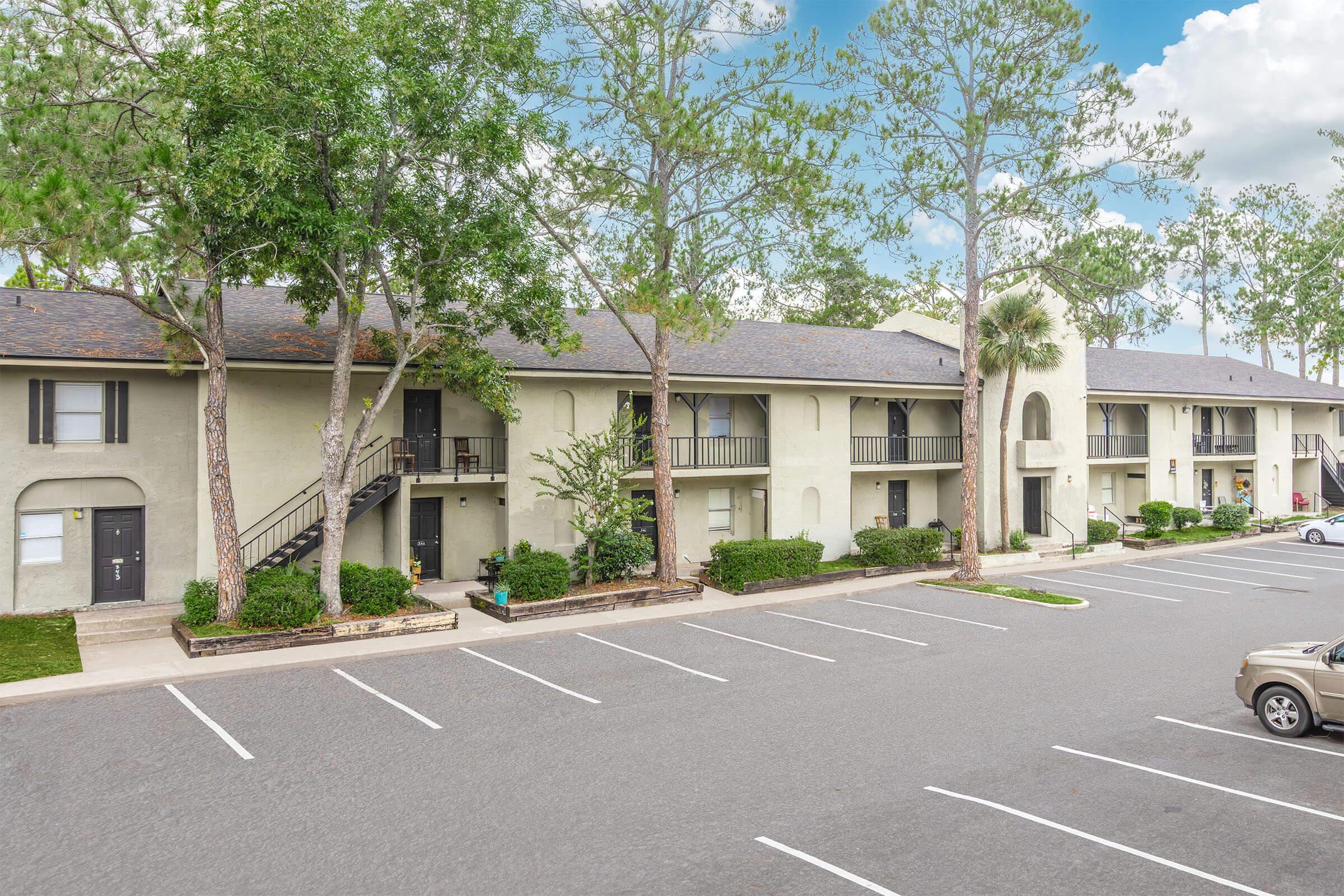 A row of multi-unit residential buildings surrounded by trees, with a small parking lot in front. The buildings feature balconies, and there are several parking spaces marked on the pavement. The scene is bright and inviting, with clear blue skies above.