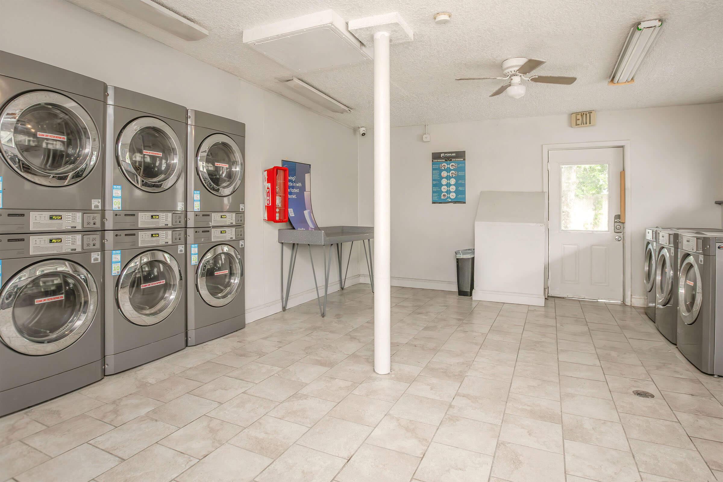 A laundromat interior featuring multiple stacked washing machines and dryers along one wall. There's a folding table in the center, a trash bin, and a door leading outside. Bright lighting and tiled floors create a clean, functional space.