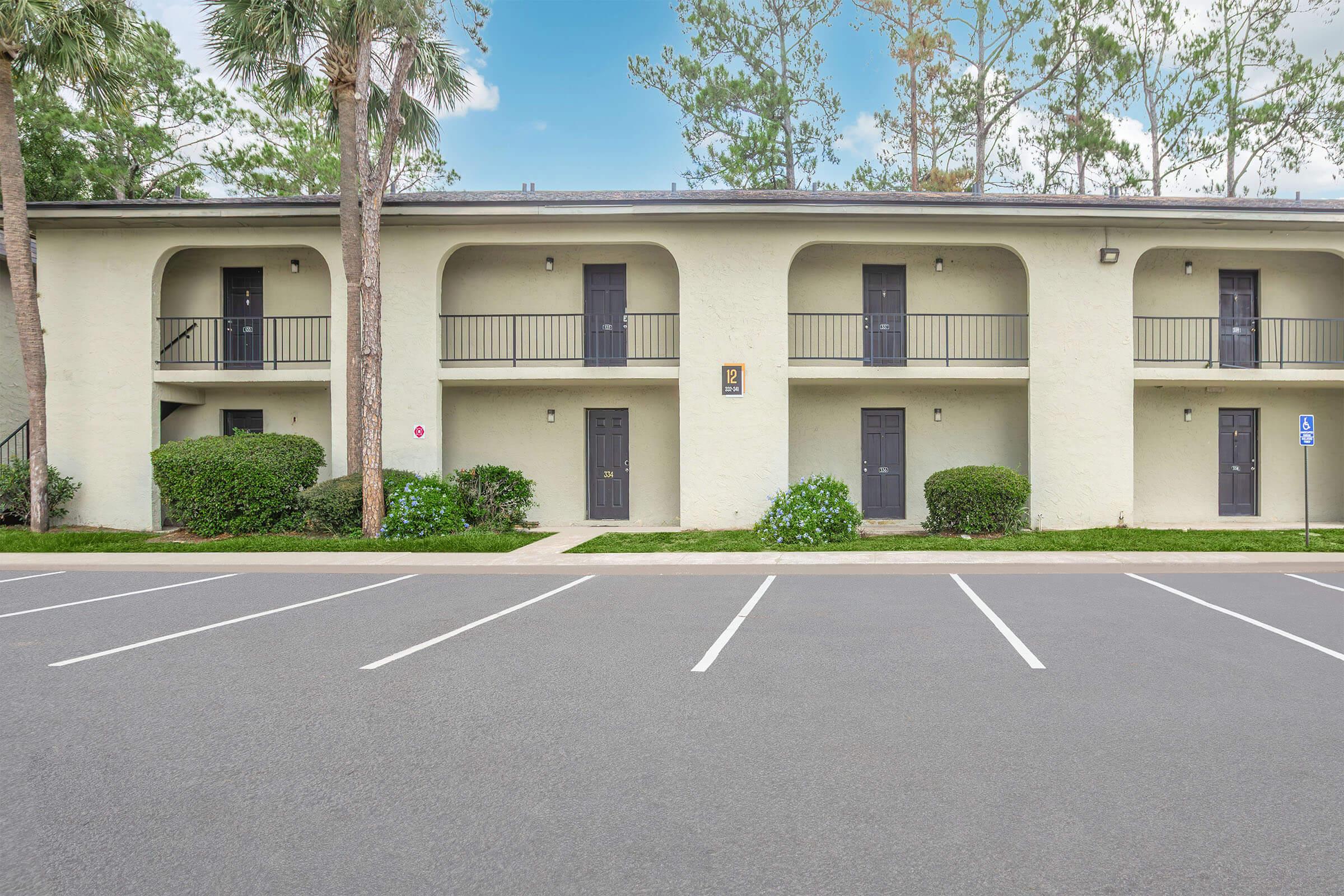 Two-story building with multiple entrances, featuring four black doors and balconies. The building is surrounded by green bushes and trees, with a clear blue sky above. A parking lot in front has several marked parking spaces.