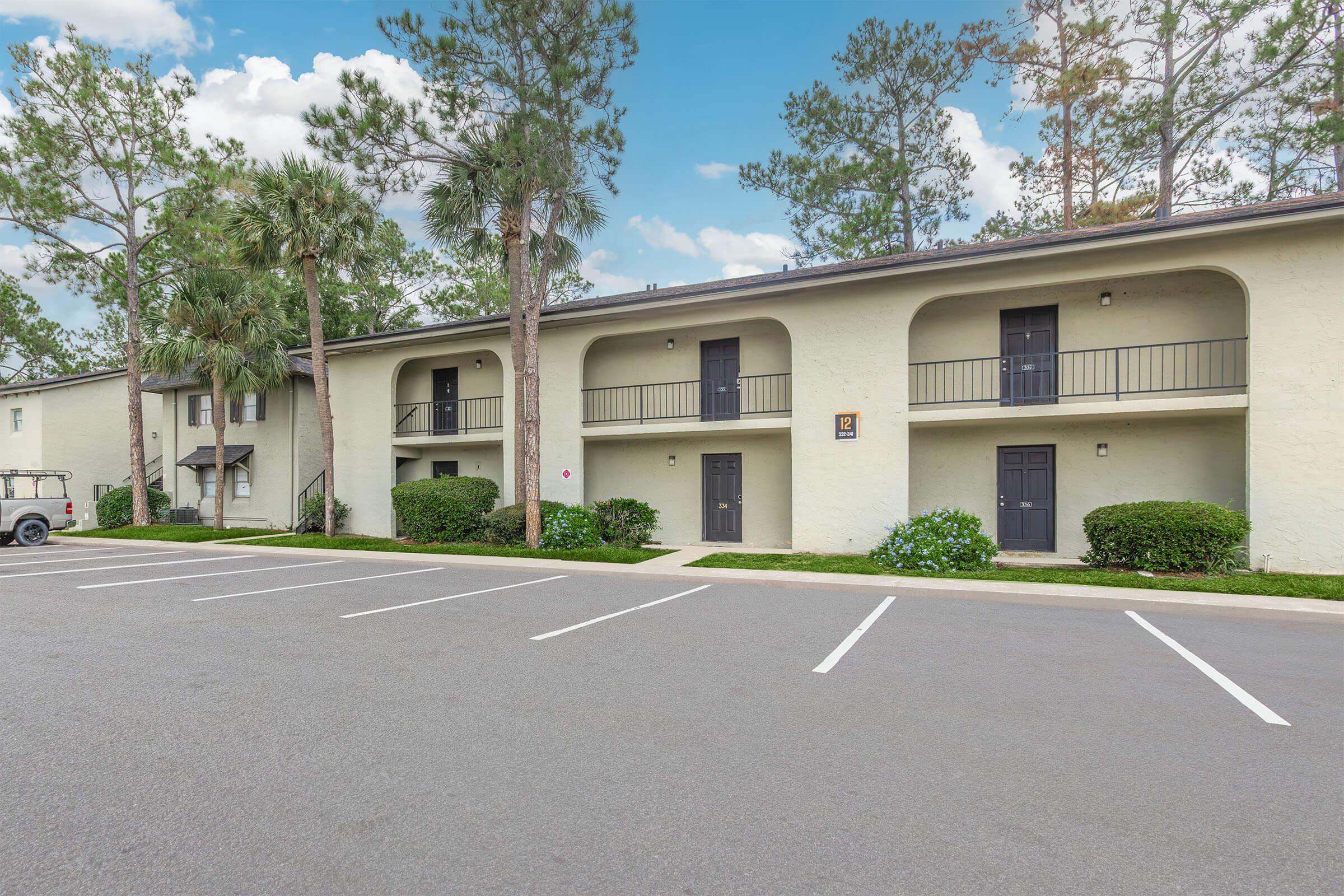 A view of a multi-story building surrounded by palm trees and greenery. The building features balconies on the upper floor and dark-colored doors. A parking lot is visible in front with marked parking spaces, and the area is set against a backdrop of blue skies and trees.