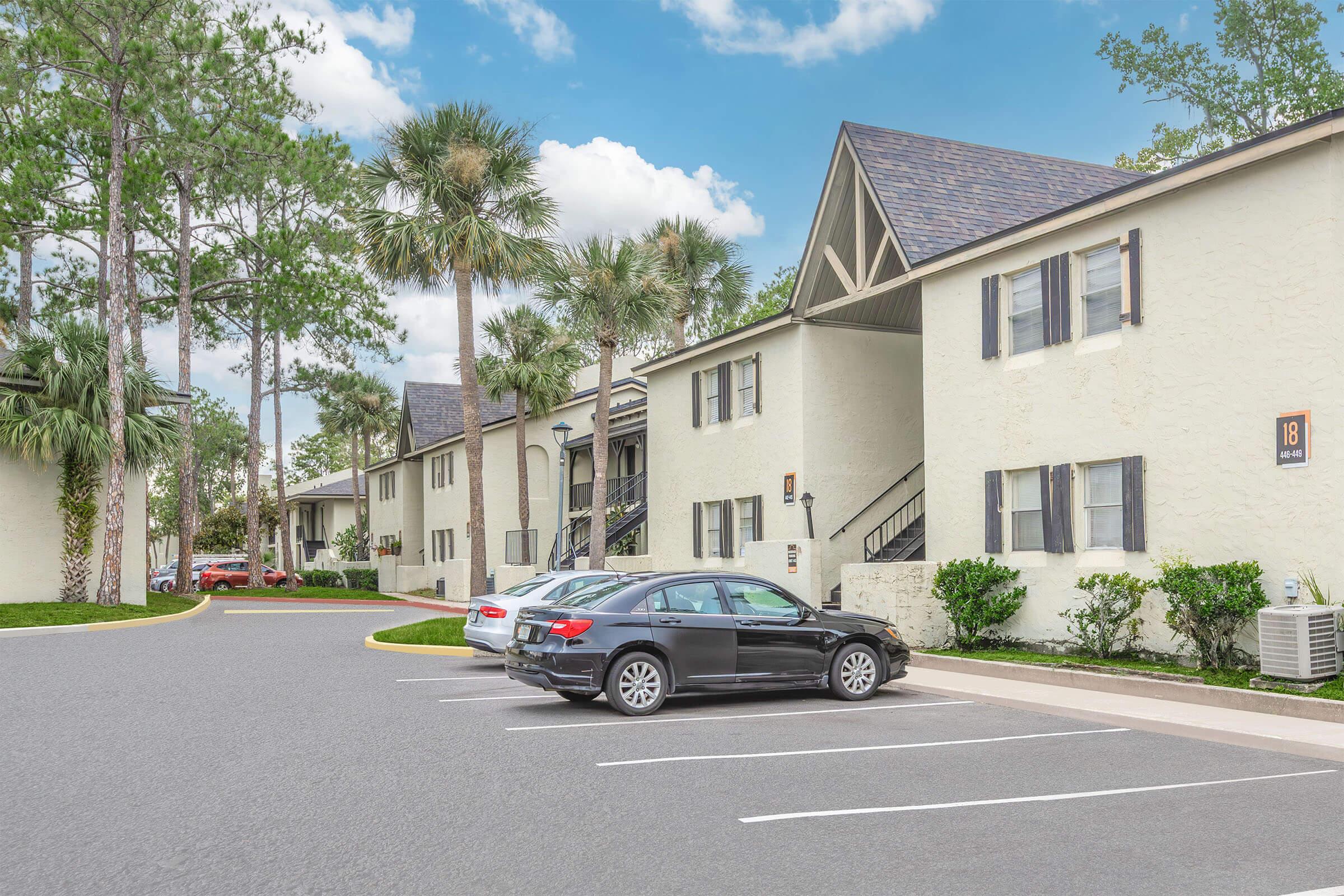 A residential complex featuring two-story buildings surrounded by trees and palm plants. The parking lot in front has several parked cars, including a black sedan. The sky is partly cloudy, creating a bright and inviting atmosphere.