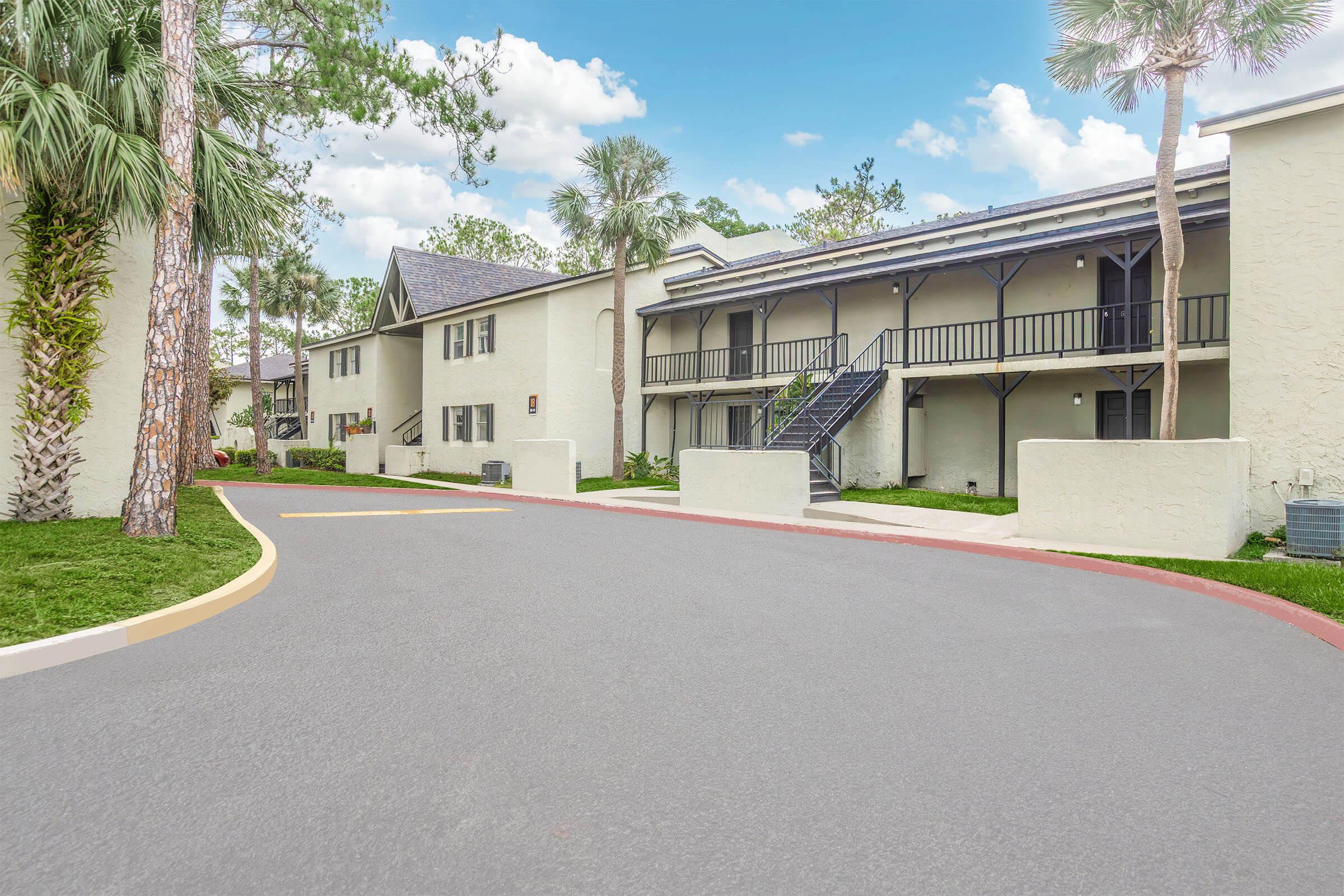A well-maintained residential complex featuring two-story buildings with balconies. The entrance drive is lined with palm trees and well-manicured landscaping. The sky is clear with scattered clouds, creating a bright and inviting atmosphere.