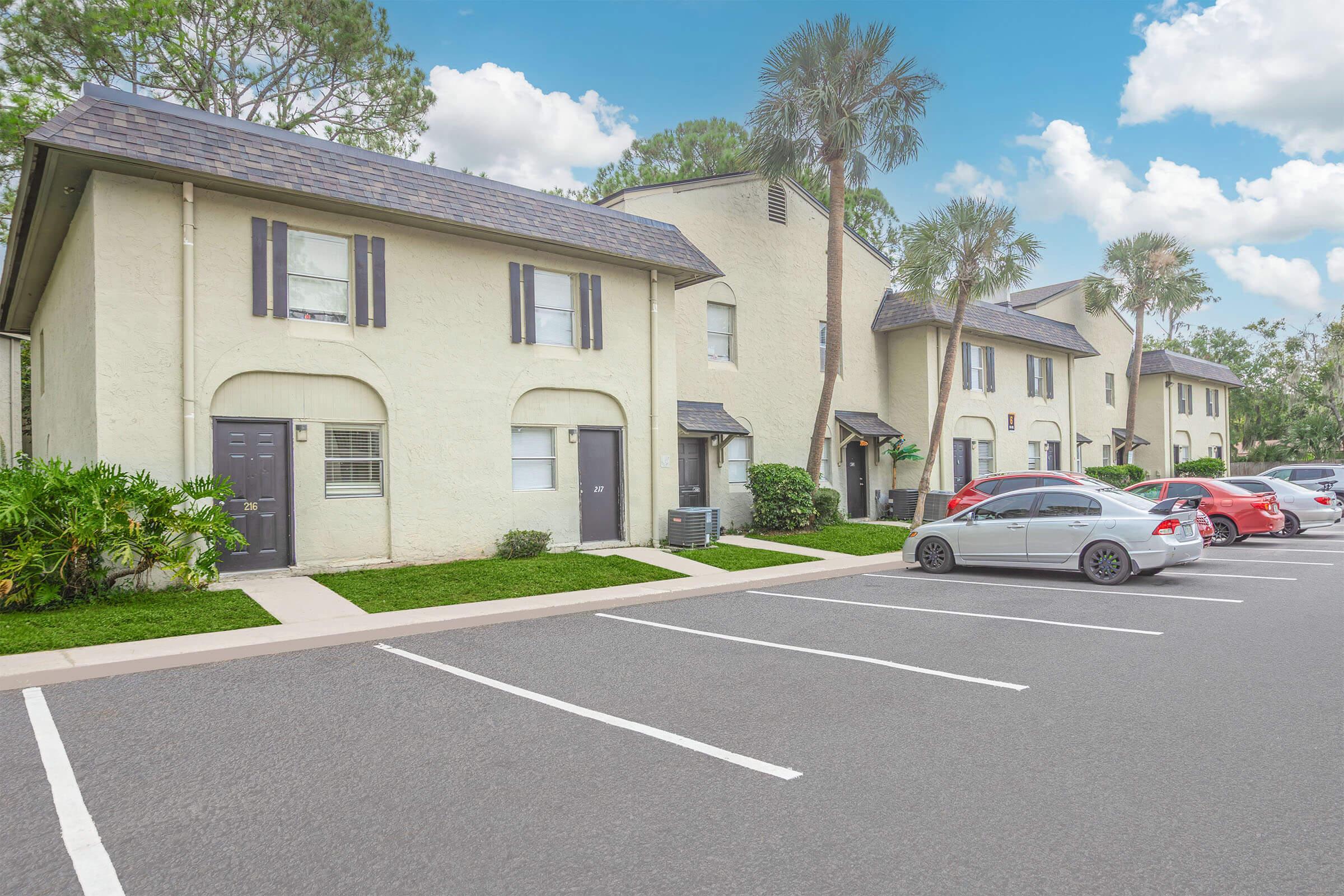 A row of beige apartment buildings with black shutters, surrounded by well-maintained landscaping and palm trees. A parking lot in the foreground has several parked cars, including a silver sedan and red vehicles. The sky is partly cloudy, adding a bright ambiance to the scene.