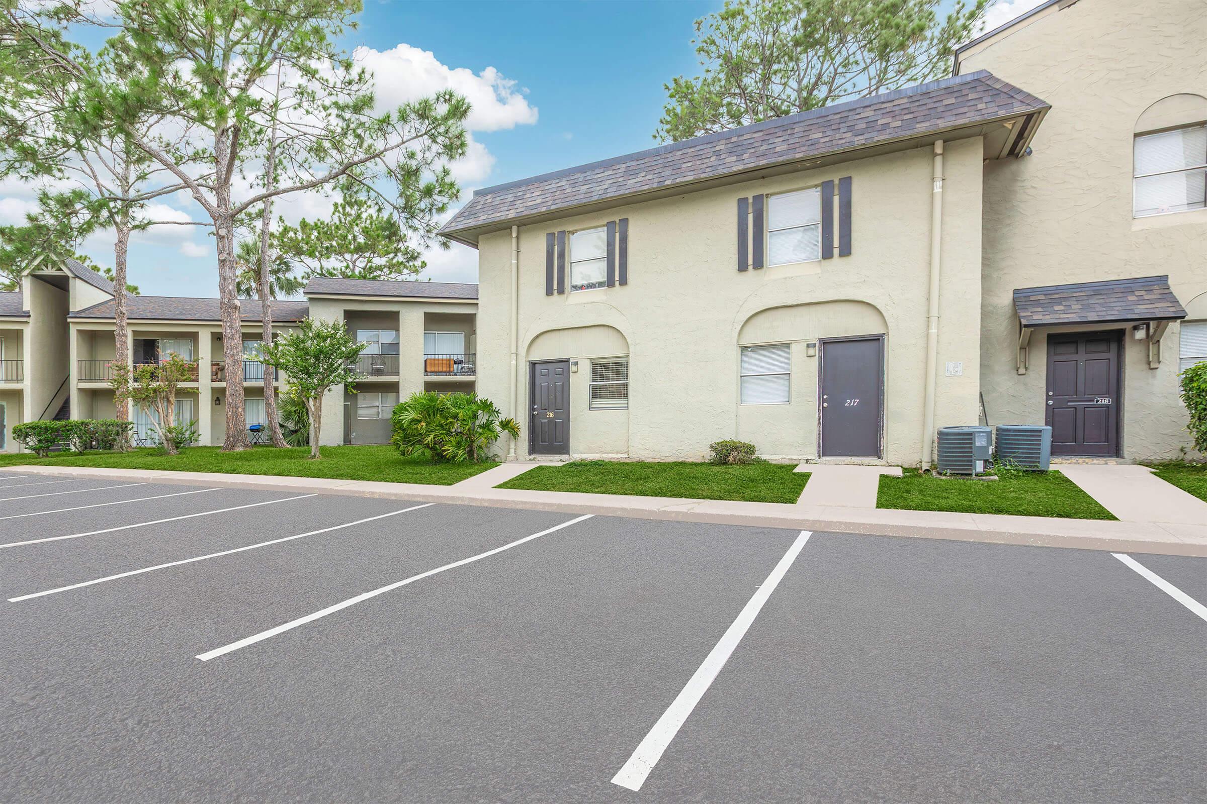 A row of apartment buildings with a well-maintained landscape. The buildings are beige with dark shutters, and each has a front door. In the foreground, there is a parking lot with several empty spaces. Lush green trees are visible in the background against a blue sky.