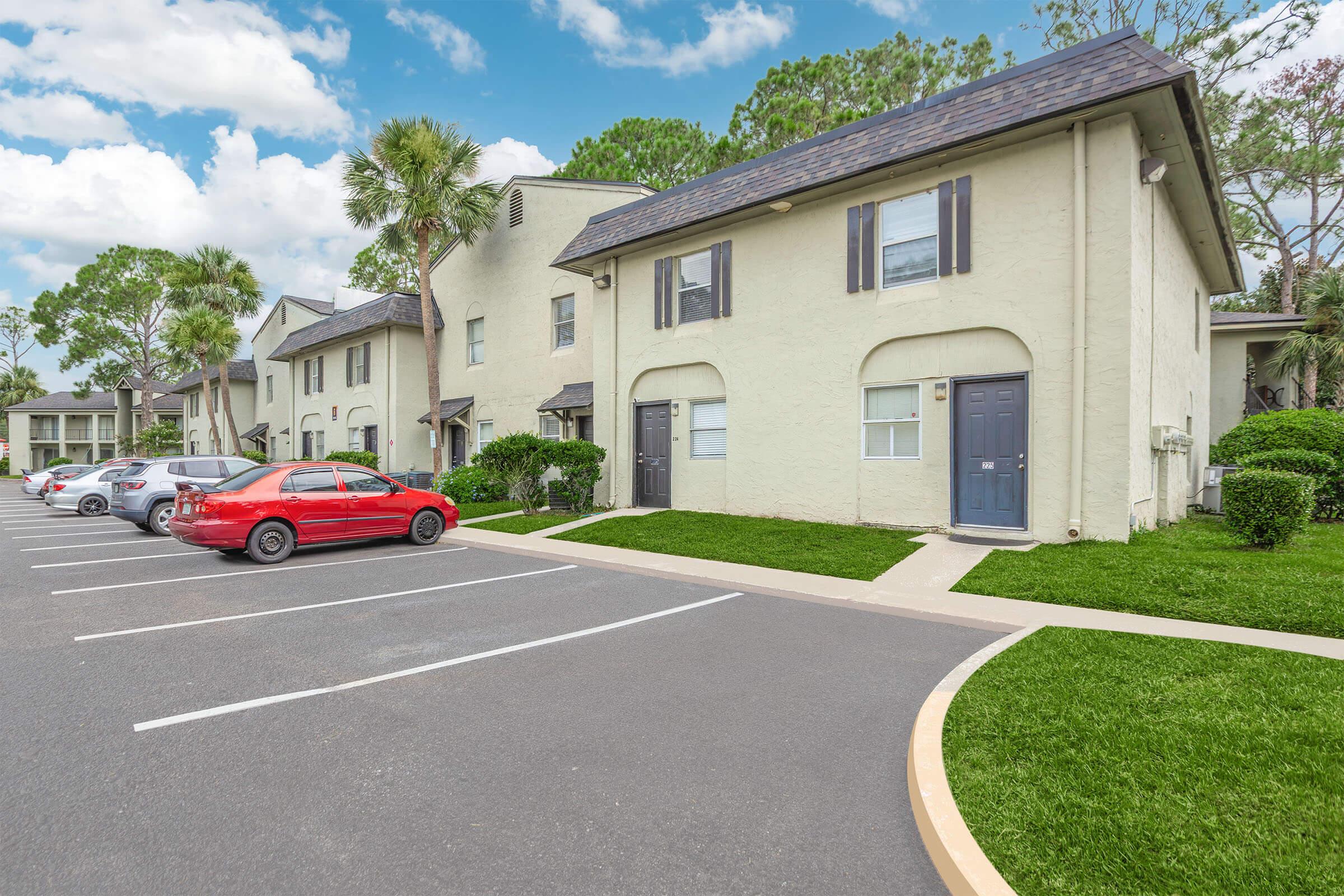 A residential area featuring several two-story apartment buildings surrounded by greenery. The image shows a parking lot with parked cars, including a red car in the foreground, and well-maintained lawns. The sky is partly cloudy, contributing to a bright and inviting atmosphere.