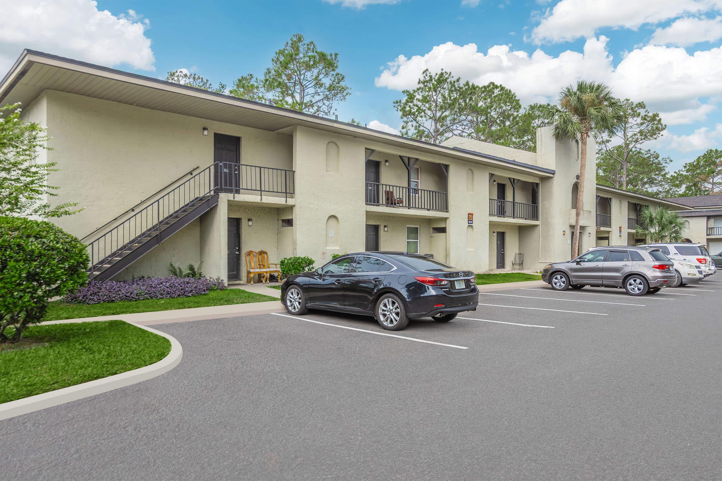 A view of a two-story apartment building with a staircase and parking area. In the foreground, there are parked cars, including a black vehicle. The surrounding area features neatly trimmed grass and shrubs, with trees in the background and a partly cloudy sky.