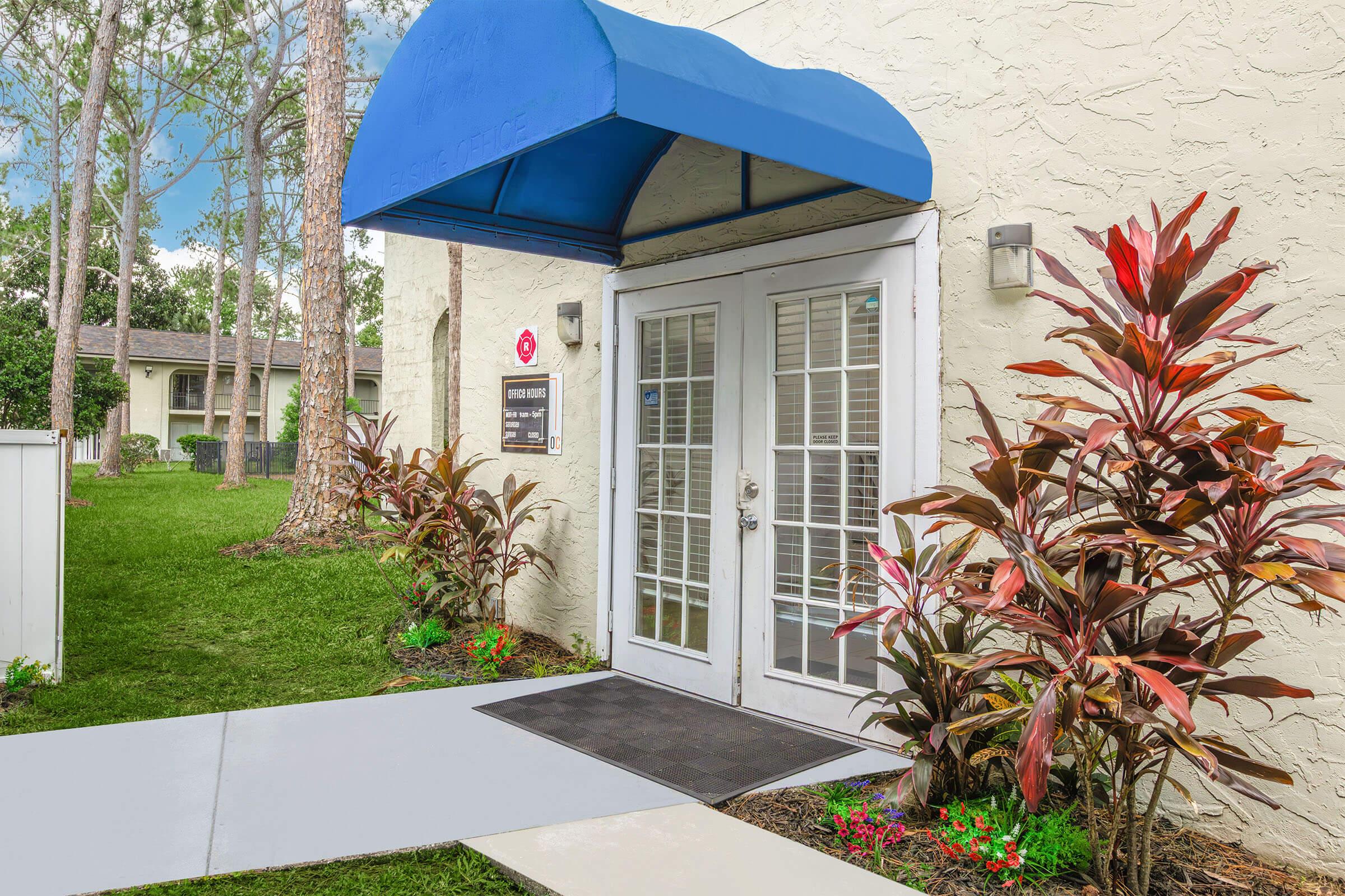 Entrance of a building featuring double glass doors with a blue awning above. The path is lined with greenery and colorful plants, including large-leaved foliage. In the background, a well-maintained grassy area and additional building can be seen, creating a welcoming atmosphere.