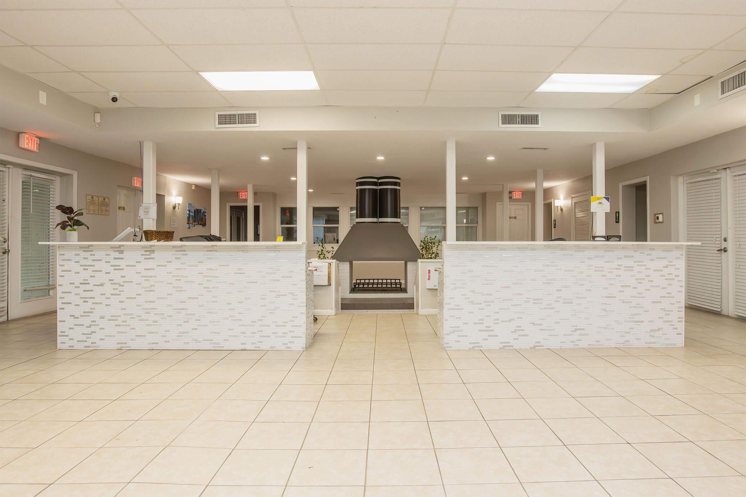 Interior view of a modern reception area featuring two white-tiled counters. The space is well-lit with overhead lights, and a large central chimney is visible. There are sliding doors on both sides leading to additional rooms, and potted plants add a touch of greenery to the setting.