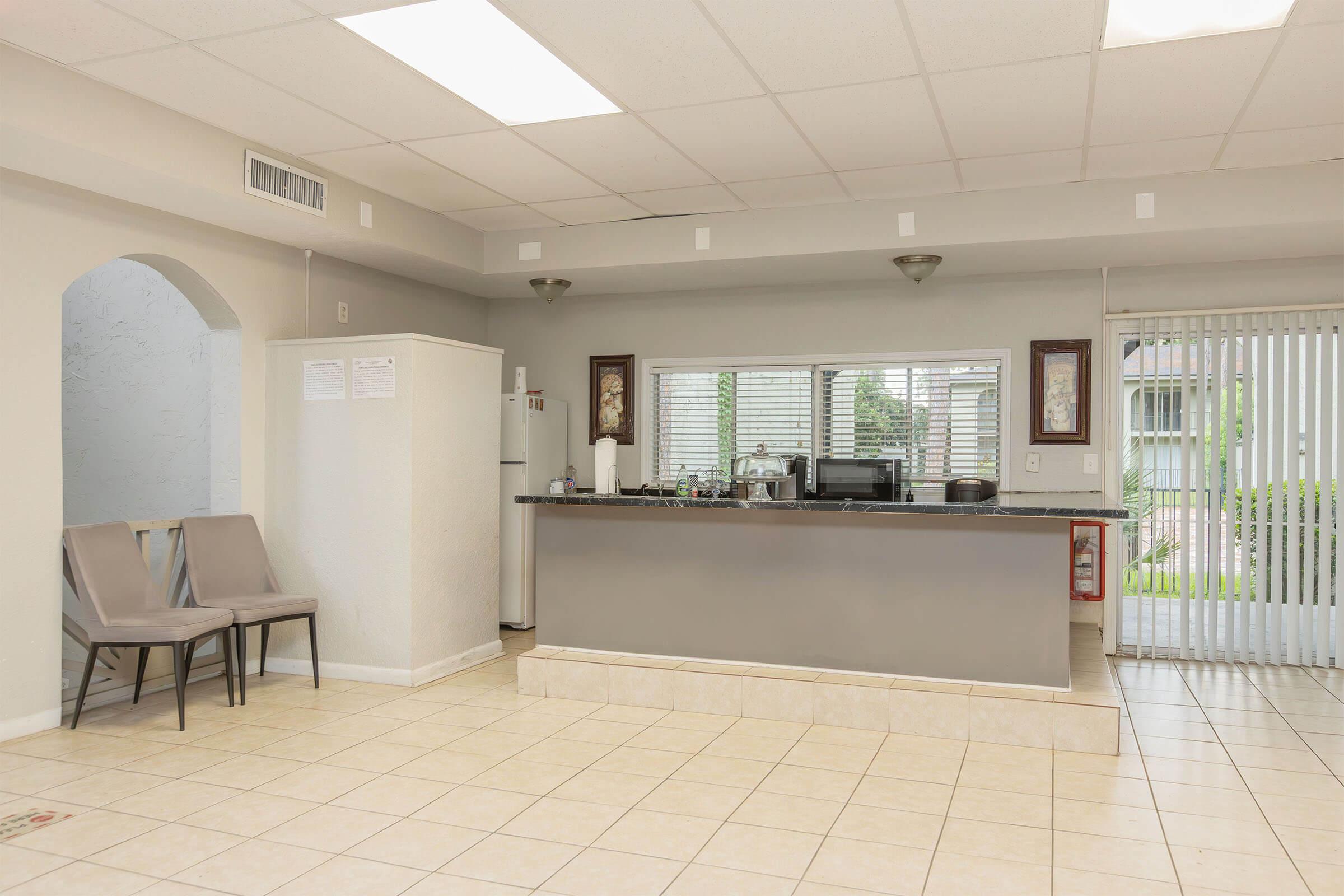 A bright, modern reception area featuring a gray counter, a coffee station, and two empty beige chairs against the wall. Large windows allow natural light to fill the space. The floor is tiled, and there are decorative elements on the walls, creating a welcoming atmosphere.