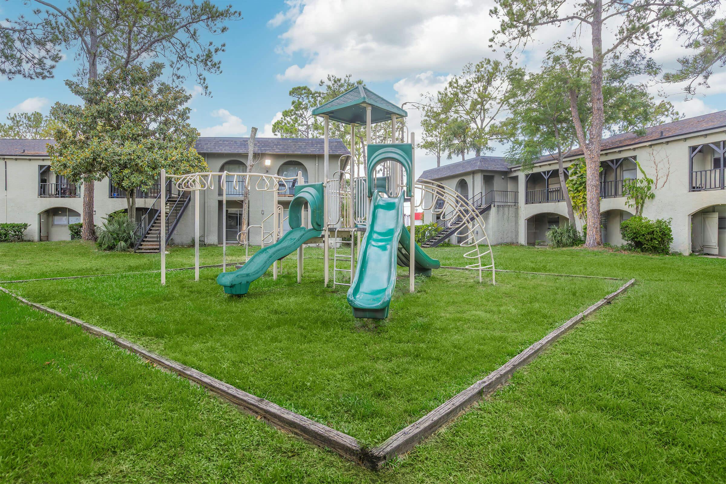 Playground featuring two green slides and climbing equipment, set on a lush green lawn. The background includes an apartment complex with balconies and trees. The sky is partly cloudy, creating a bright and inviting atmosphere for outdoor play.