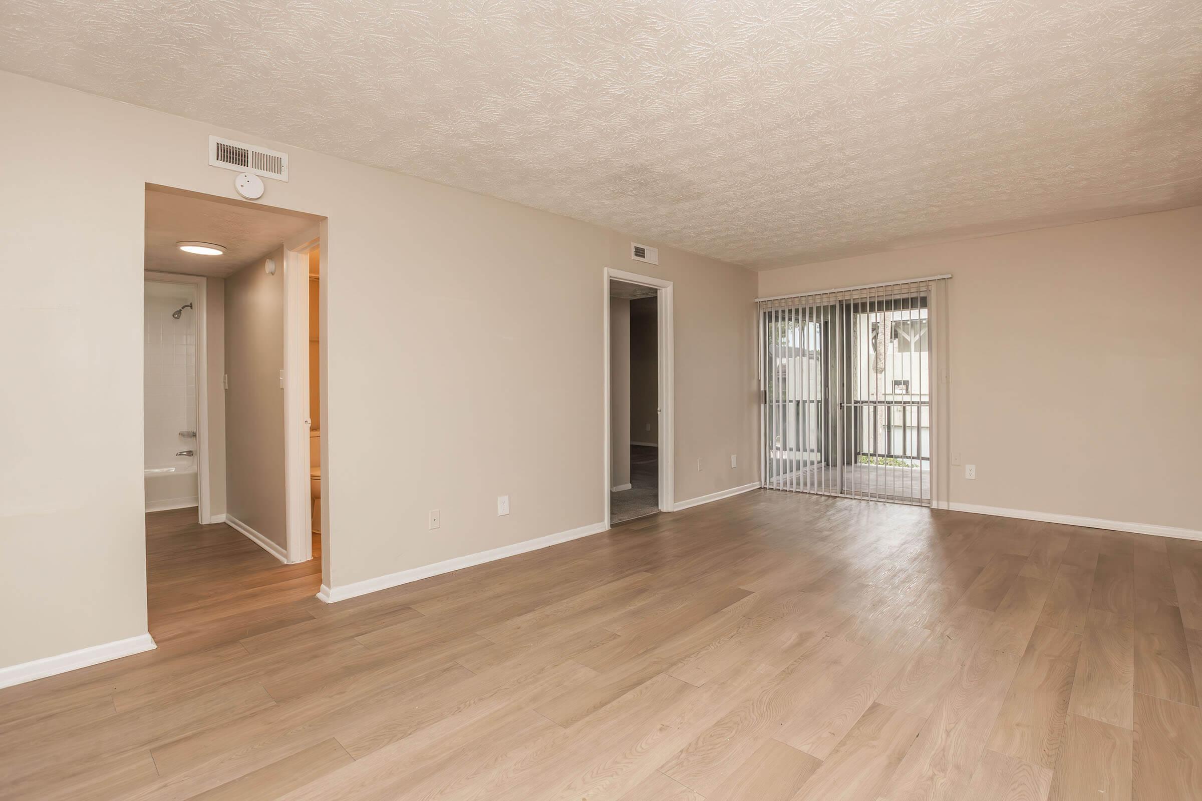 A spacious, empty living room with light-colored walls and wooden flooring. There is a sliding glass door leading to a balcony on the right, and an entrance to a bathroom on the left. The room is well-lit with natural light, creating a welcoming atmosphere.