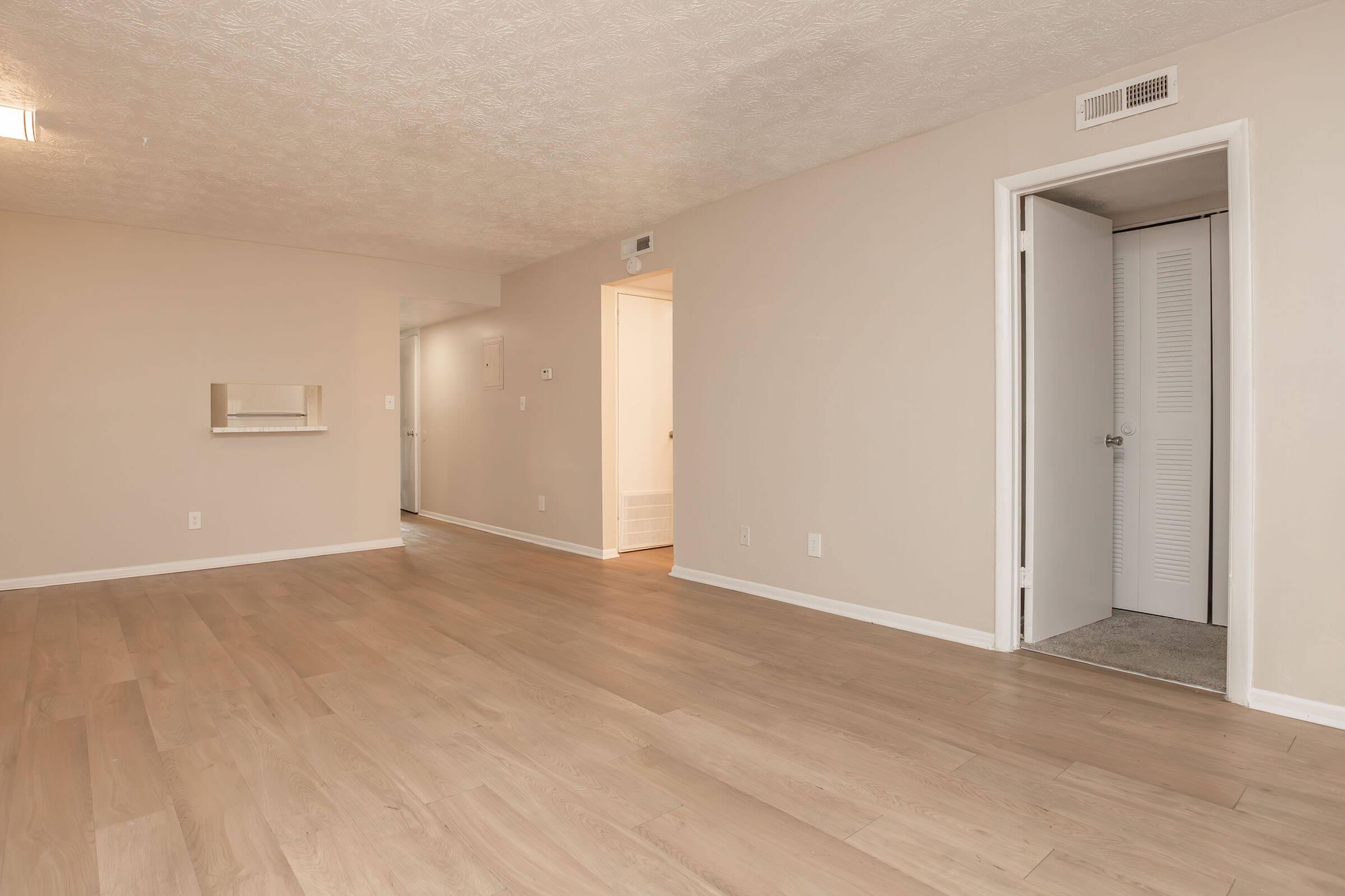 A spacious, empty living area with light-colored wooden flooring. The walls are painted in a soft beige hue, creating a warm ambiance. A small opening for a wall-mounted shelf is visible on the left, while a doorway leads to another room on the right. Natural light brightens the area from a ceiling fixture.