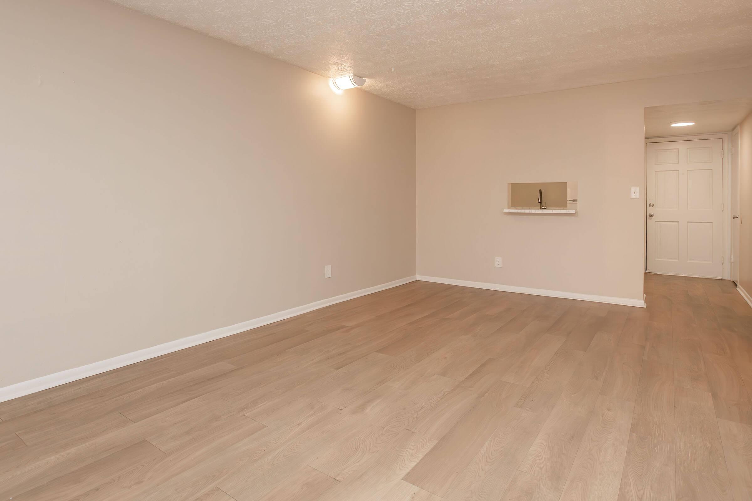 Interior view of a spacious, empty room with light-colored wooden flooring and neutral beige walls. A small cut-out wall shelf is visible on one side, and there's a doorway leading to another room in the background. Soft overhead lighting brightens the space, giving it a clean and modern appearance.