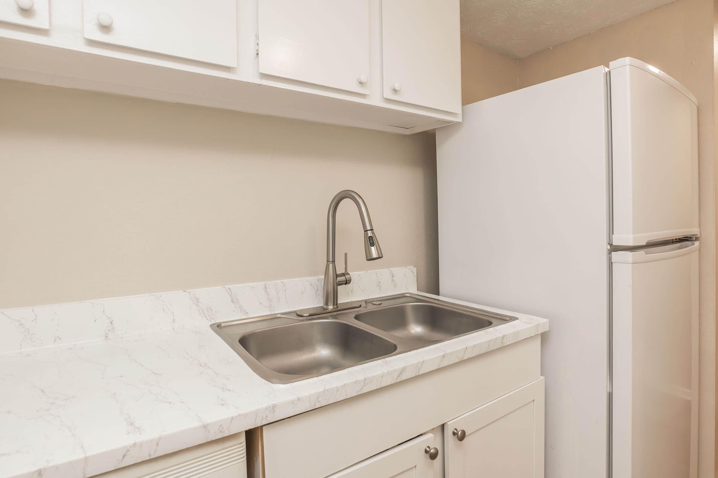 A clean and modern kitchen space featuring a white countertop with a dual sink, a sleek faucet, and white cabinetry. In the background, a white refrigerator is visible, set against a neutral wall, creating a bright and organized atmosphere.