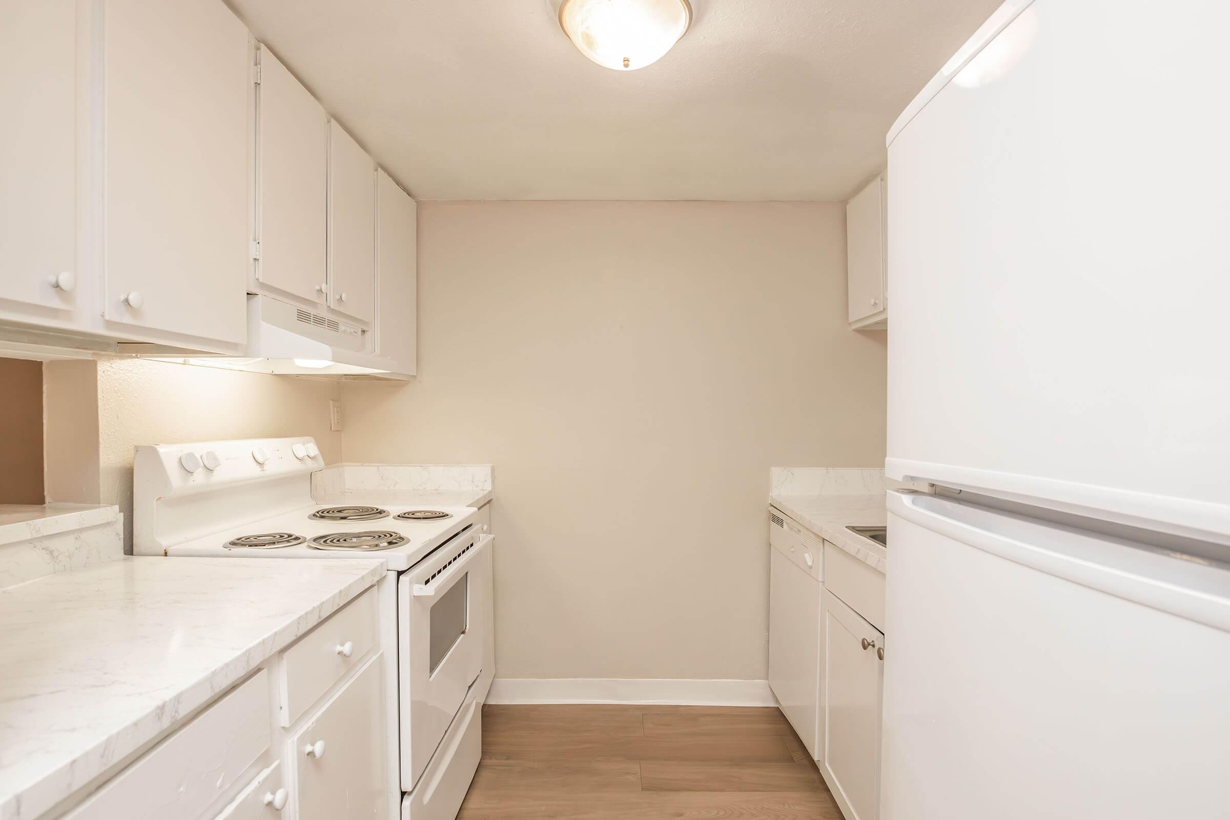 A clean and modern kitchen featuring white cabinetry, a white refrigerator, a white stove with an oven, and a countertop with a marble finish. The walls are painted a light color, and the floor has a wood-like appearance. The space is well-lit with overhead lighting.