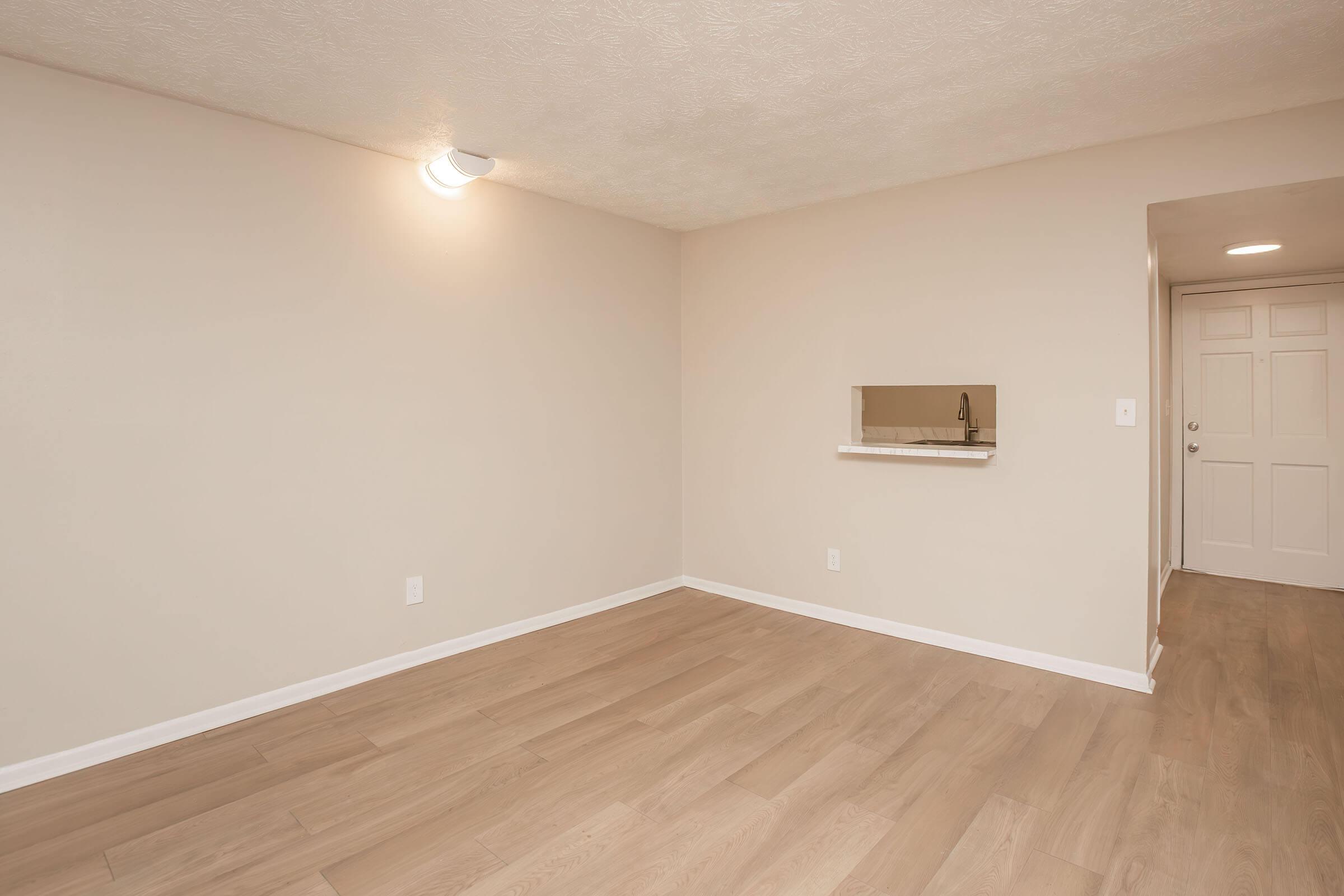 A vacant room with light-colored walls and laminate flooring. There is an opening in the wall that likely leads to a kitchen area. A door is visible on one side, and a ceiling light provides illumination. The space appears clean and ready for furniture.