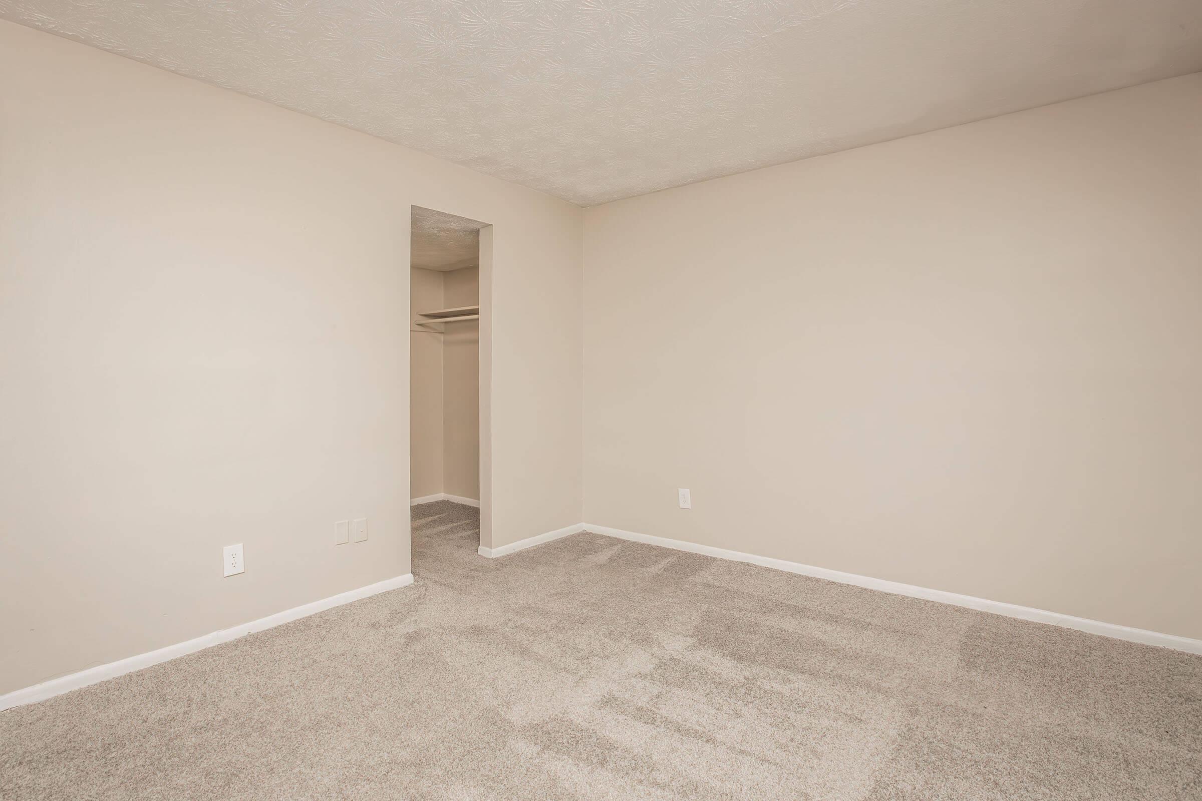 A vacant room with light-colored walls and carpet. There is a small closet visible through an open doorway on the left side. The space is well-lit and has a simple, minimalist design, creating a neutral atmosphere suitable for various purposes.