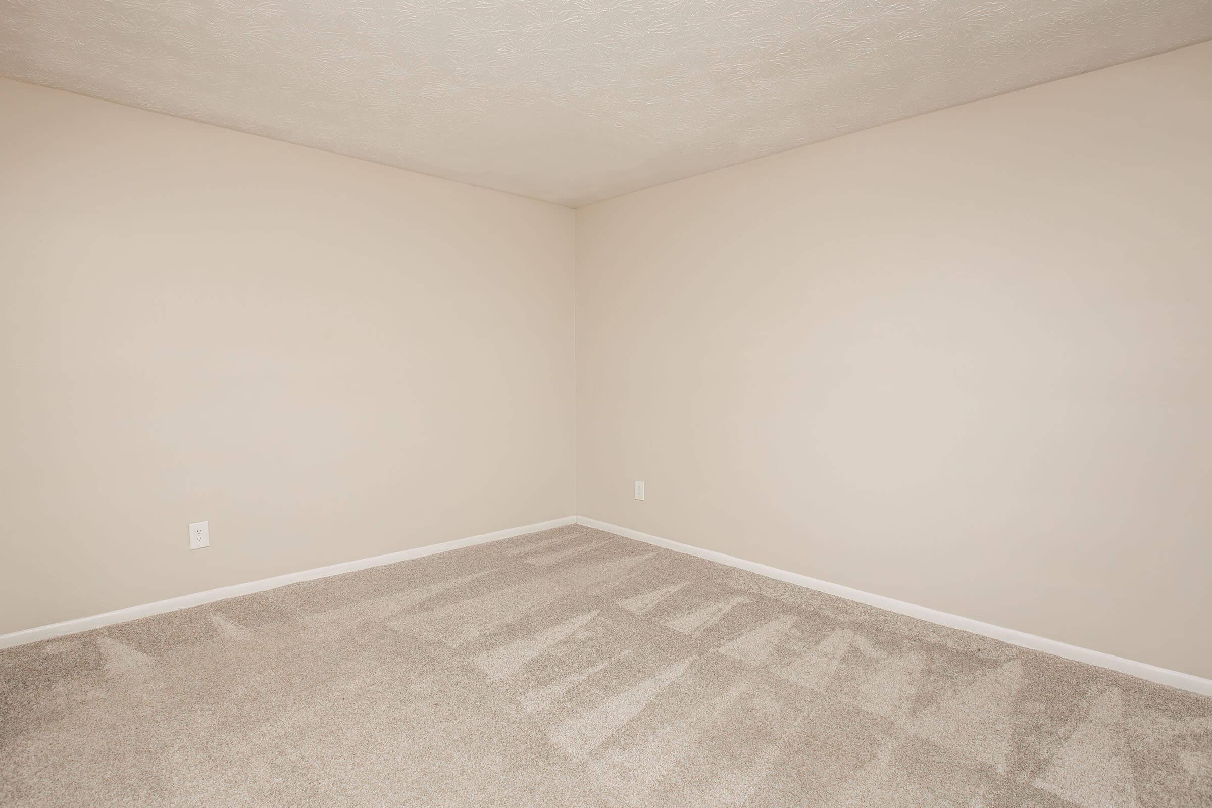 Empty room with light beige walls and a carpeted floor. The carpet shows faint indentations from furniture, and there are no windows or furnishings present. The overall atmosphere is clean and minimalistic, with plenty of open space.