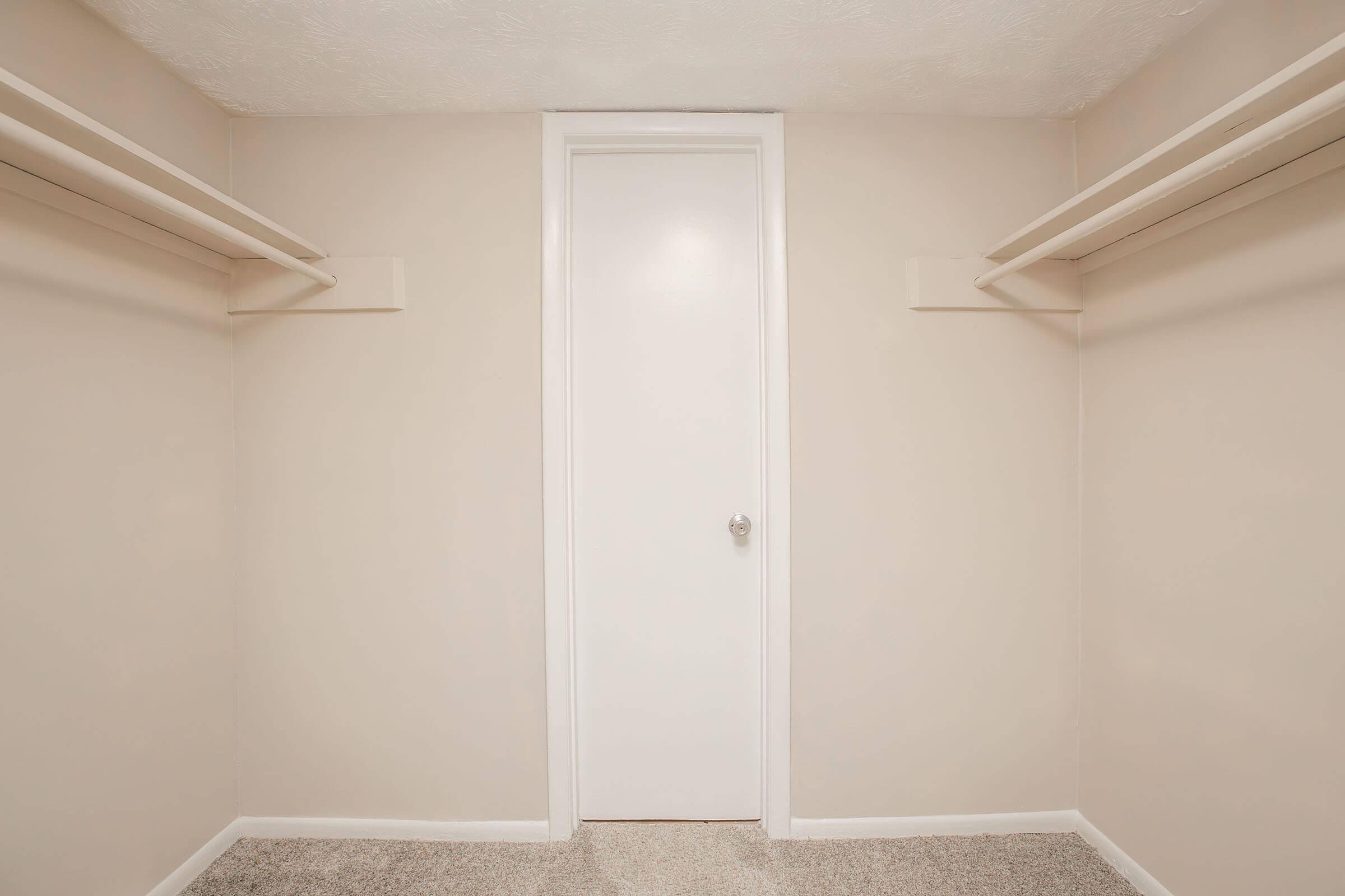 A view of an empty closet with beige walls and two shelving units on either side. The closet features a closed white door at the back, and the floor is covered in light-colored carpet. The overall appearance is clean and well-lit, suggesting a minimalist and organized space.