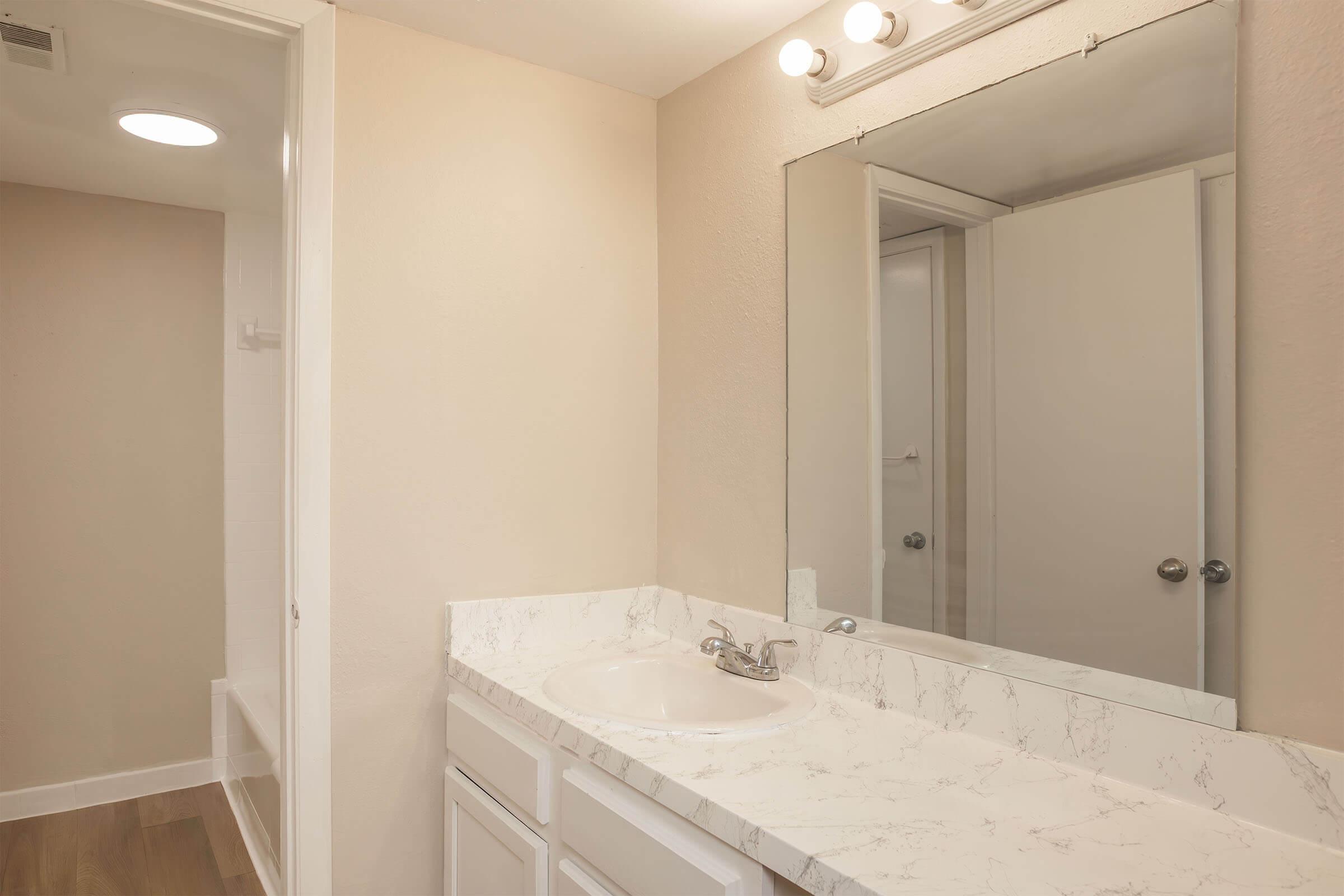 A clean bathroom with a marble countertop sink, a large mirror above the sink, and bright lighting. The walls are a soft beige color, and there is a doorway leading to a separate area, possibly a closet or restroom. The floor has a warm, wooden appearance.