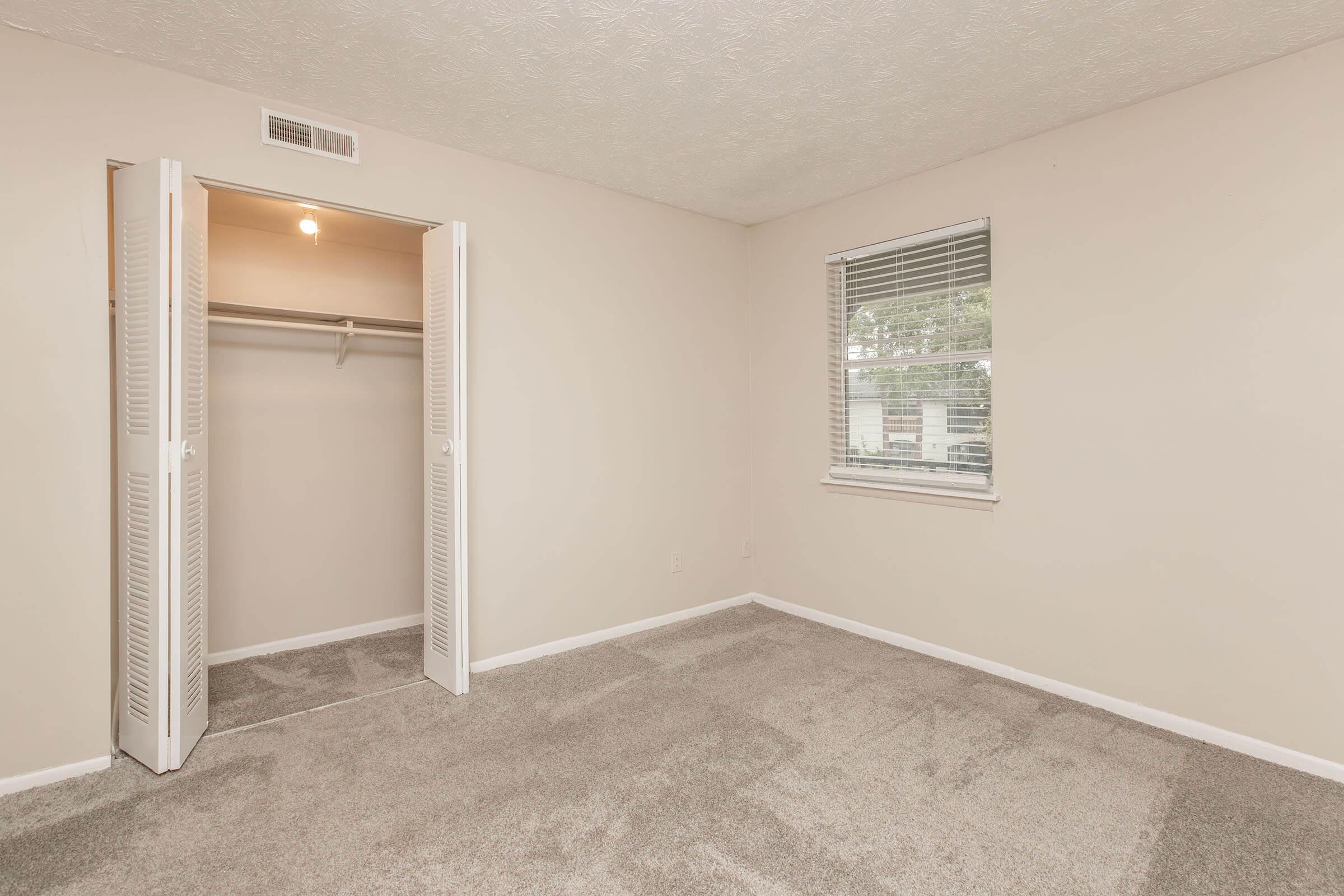 A vacant room with light-colored walls and new carpet. There's a closet with louvered doors on the left side, and a window on the right providing natural light. The space is unfurnished, creating an open and airy feel, suitable for personalized decoration.