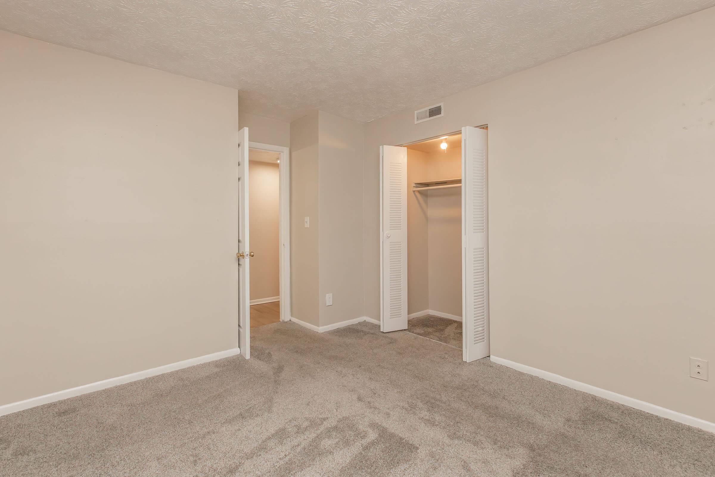 Empty room with light beige walls and soft, plush gray carpet. There is a closet with bi-fold doors on the right, and an open door leading to another room on the left. The space is well-lit, showcasing a clean and simple layout suitable for decorating.