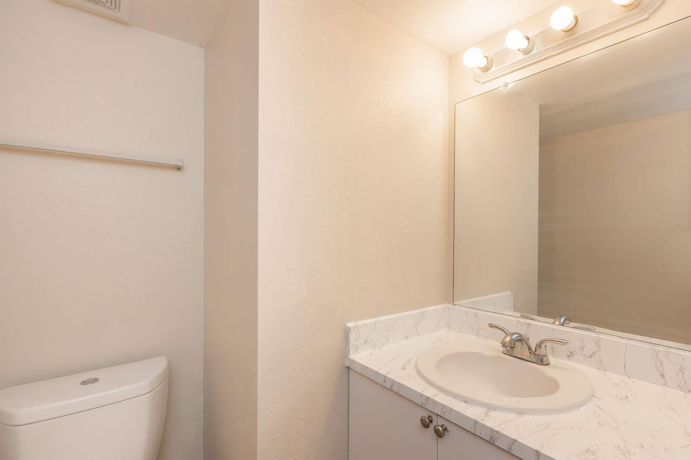 A modern bathroom featuring a white toilet, a marble countertop with a sink, and a large mirror above. There are light fixtures above the mirror, and the walls are painted in a light color, enhancing the bright and clean aesthetic of the space.