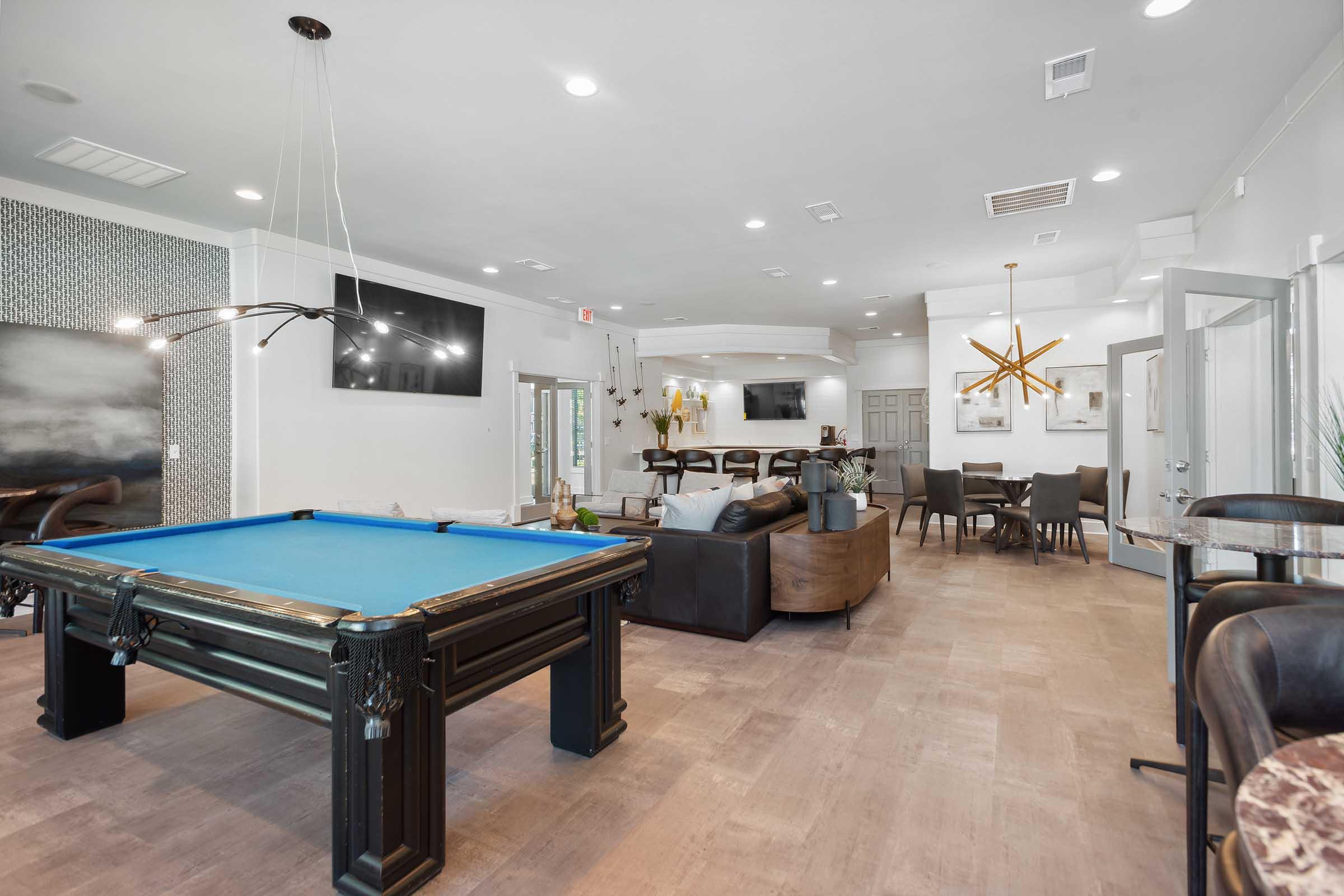 Modern common area with a pool table, comfortable seating, and a chandelier. Two flat-screen TVs are mounted on the walls. The space features neutral decor, large windows for natural light, and tables set for dining or gathering. A welcoming atmosphere for socializing and relaxation.